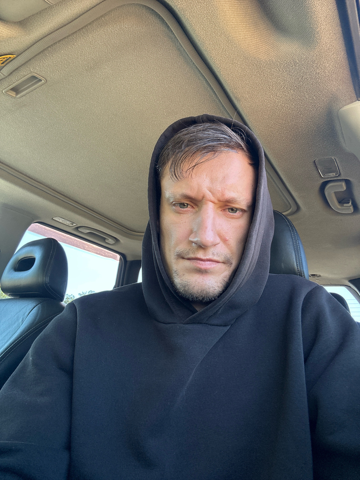 A man is sitting in the driver's seat of a car, wearing a black hoodie. The car interior is visible, including the headrest of the passenger seat, the car ceiling, and a portion of the exterior through the window. The man is looking directly at the camera with a serious expression. The background outside the window suggests it might be daytime, with a glimpse of trees and a building. The man's hair is dark and appears to be styled upwards. He has stubble on his face. The overall scene is a candid selfie taken from inside the vehicle.
