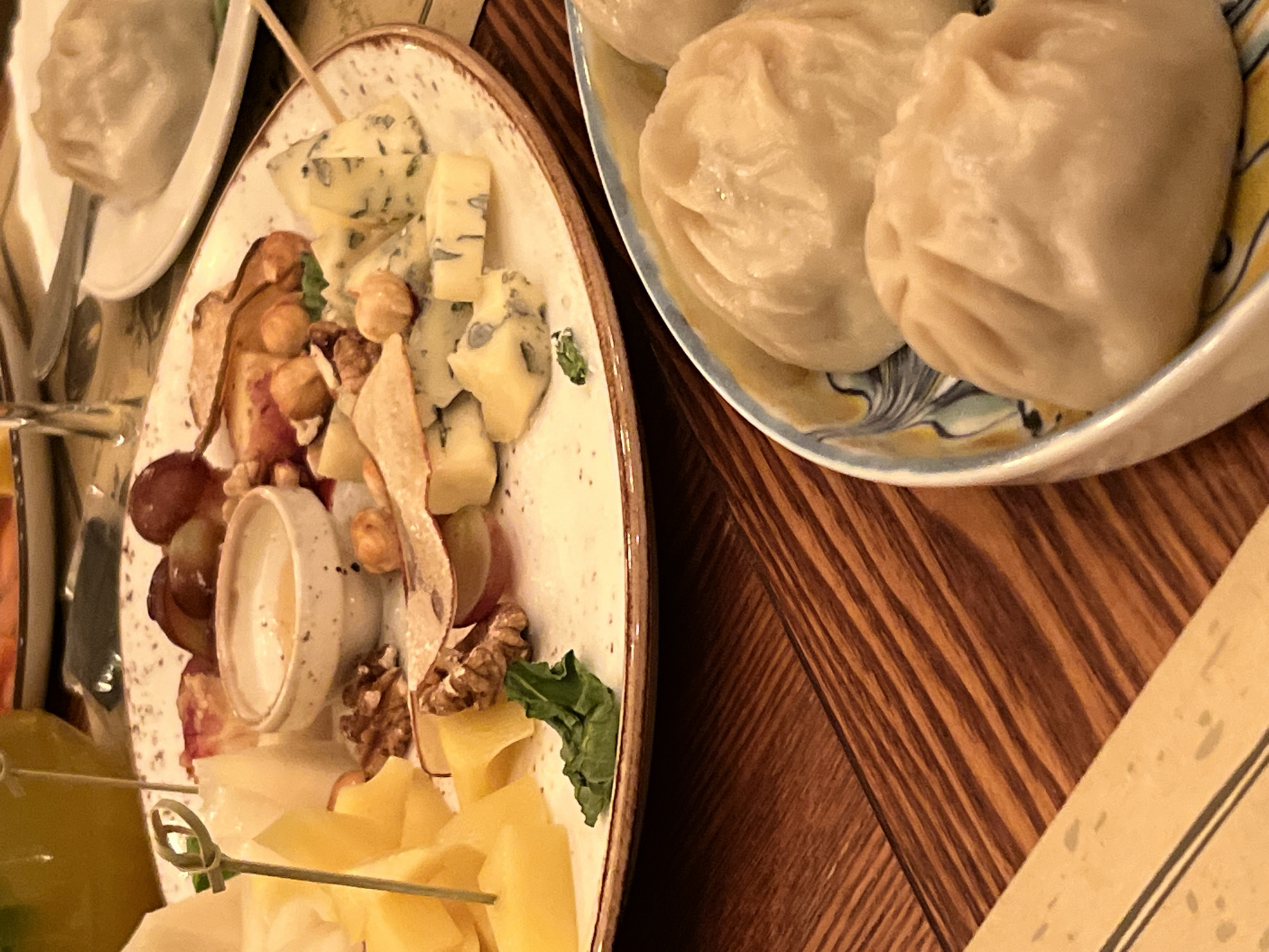 A close-up, high-angle shot shows a cheese board and a plate of dumplings on a wooden table. The cheese board is white with a brown rim and is filled with various types of cheese, grapes, nuts, and a small bowl of honey. The dumplings are light-colored and arranged in a bowl with a decorative pattern. The wooden table has a distinct grain pattern. The image is lit from above and to the left, casting subtle shadows. The background is out of focus but suggests a restaurant setting with other tables and a yellow drink. The scene appears to be in Novosibirsk, Russia, based on the context provided.