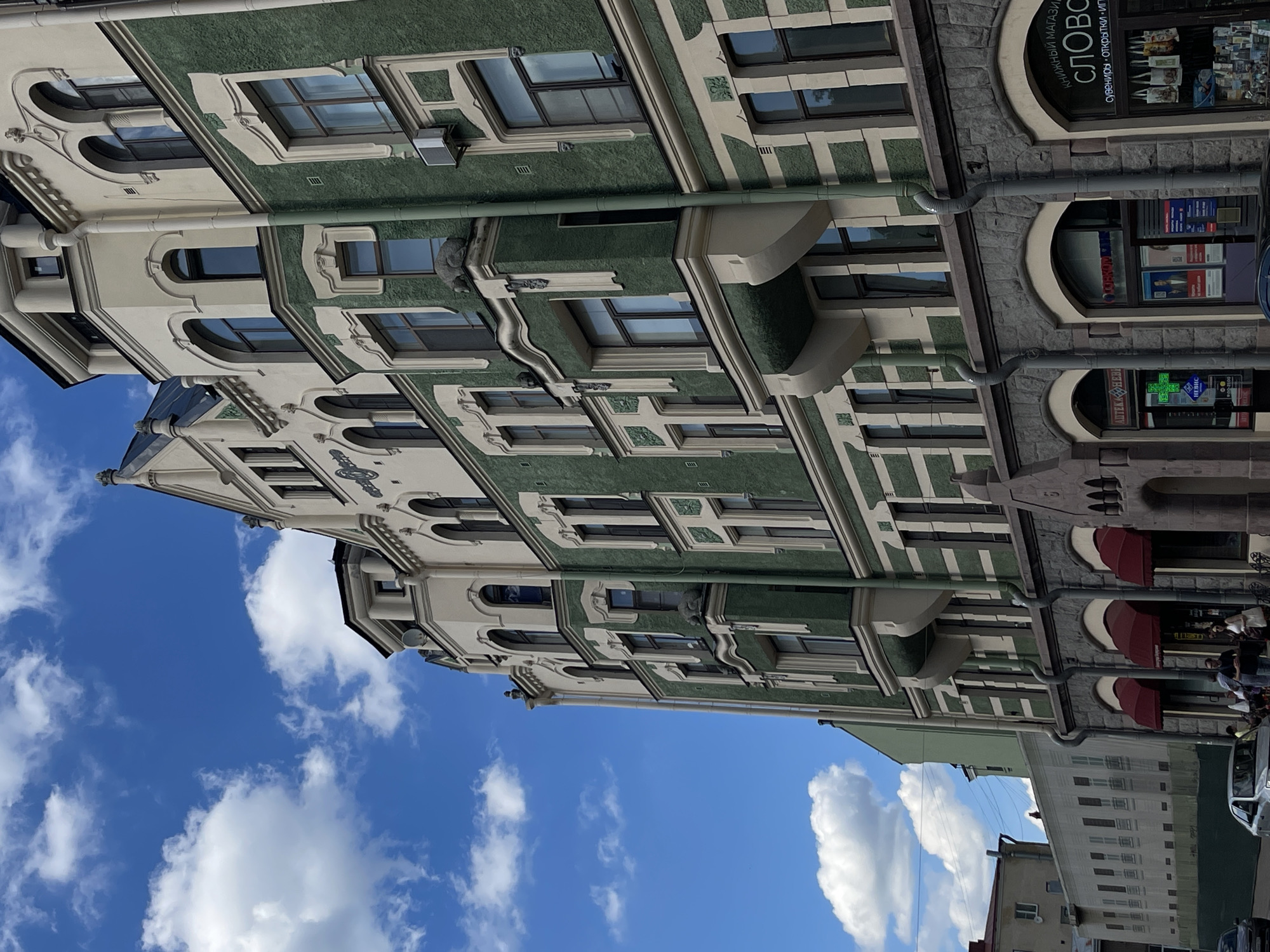 The image captures a multi-story building with distinctive green and cream architectural details, characteristic of Art Nouveau style. The building features numerous arched windows, decorative stone carvings, and prominent drainpipes running down its facade. The sky above is a vibrant blue, dotted with fluffy white clouds, suggesting fair weather. At street level, several storefronts are visible, including a pharmacy with a green neon cross and a bookstore with merchandise displayed in the window. A few people are seen walking on the sidewalk and near the entrance of the shops. Cars are parked along the street. The building is identified as being located in the city of Vyborg, Russia. The overall scene depicts a well-preserved historical urban environment.