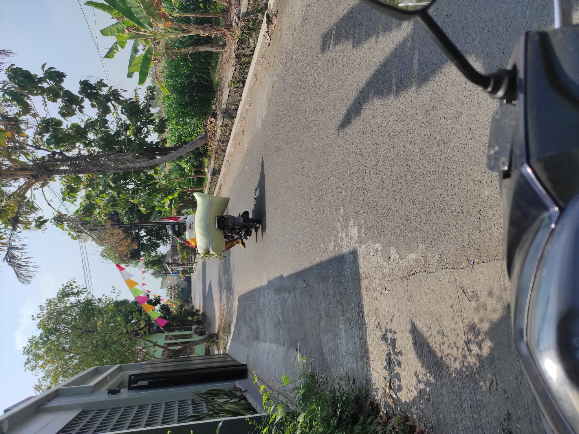 In Pleret, Indonesia, a person on a motorcycle is traveling down a paved road. The motorcycle carries a large, light-colored sack on its rear. The rider is wearing a helmet and appears to be wearing a yellow shirt and brown pants. The road is bordered by a concrete sidewalk on the left and a drainage ditch and vegetation on the right. Several houses and trees line the street, and colorful triangular flags are strung across the road in the distance, suggesting a local celebration or event. The sky is clear and blue, and the sun casts shadows on the road and sidewalk. The perspective of the photo is from another motorcycle, with its handlebar and dashboard visible in the foreground.