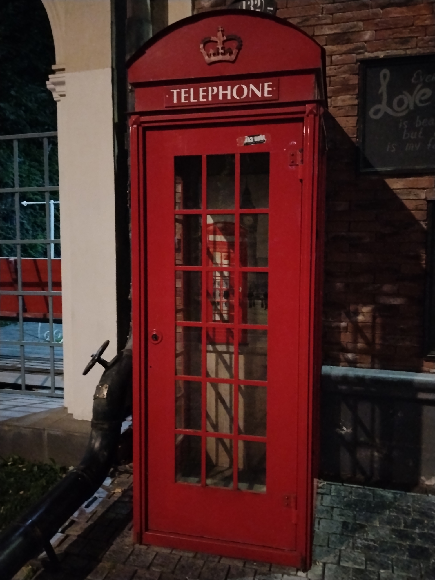 A bright red, classic British telephone booth stands prominently in an outdoor setting at night. The booth is adorned with a golden crown emblem at the top and the word "TELEPHONE" written in white letters on a black background above the glass-paneled door. A small sticker, partially obscured, is visible on the door. Inside the booth, a reflection of another red telephone booth and some interior details can be seen through the glass.
To the left of the booth, an arched structure with white columns is partially visible, and beyond that, a metal fence encloses some greenery. A dark, industrial-looking pipe with a wheel-like handle extends from the ground near the telephone booth.
To the right of the booth, a brick wall serves as a backdrop. A chalkboard with white lettering, appearing to be a quote or message about "Love," is mounted on the brick wall. The lighting suggests it is evening or night, and the scene is likely in a public or commercial area, given the presence of a telephone booth and decorative elements. The ground is paved with cobblestones. The overall mood is somewhat whimsical, evoking a sense of nostalgia or a thematic decorative element. No people are visible in the image. The location is identified as Bishkek City, Kyrgyzstan.