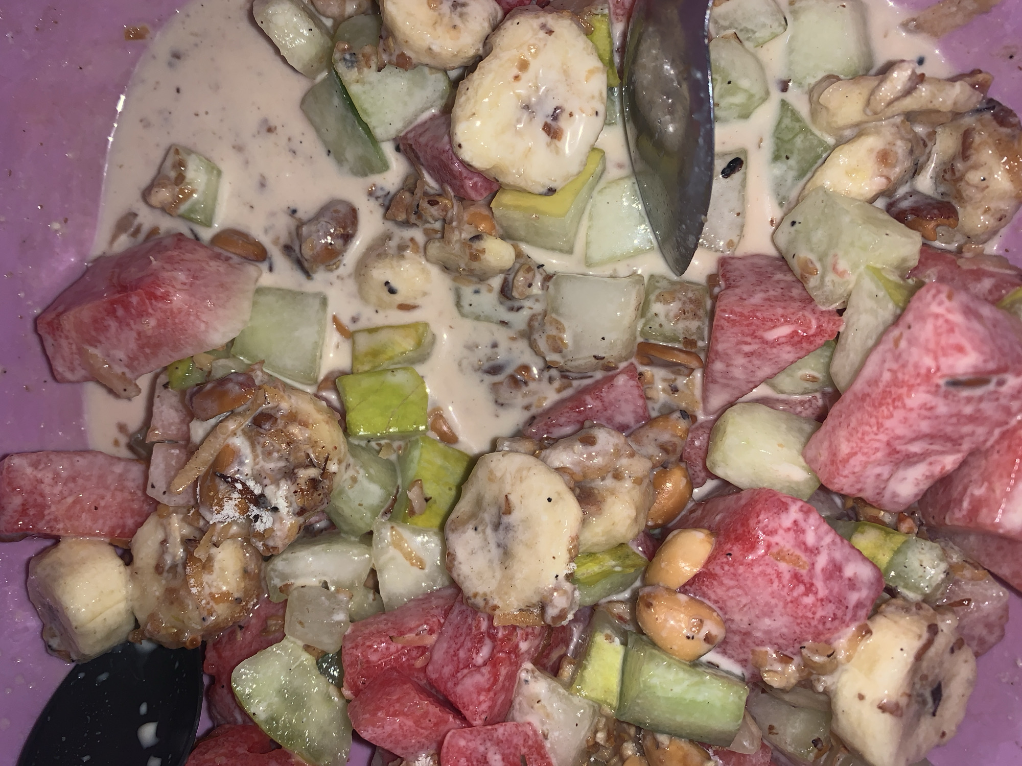 The image displays an overhead, close-up view of a vibrant fruit salad served in a purple bowl. The salad is composed of various diced fruits, prominently featuring red watermelon cubes, sliced yellow bananas, and greenish-white cubes, likely cucumber or honeydew melon. These fruits are coated in a creamy, off-white dressing or yogurt. Small, light brown nuts, possibly peanuts, are interspersed throughout the mixture, adding textural variety. A metallic spoon is partially visible on the right side of the bowl, suggesting the dish is prepared for eating. The scene focuses entirely on the food, showcasing its colorful ingredients and creamy consistency. No text is visible in the image, and while the provided context indicates Rafin Par, Nigeria, there are no visual cues within the image to confirm this specific location.