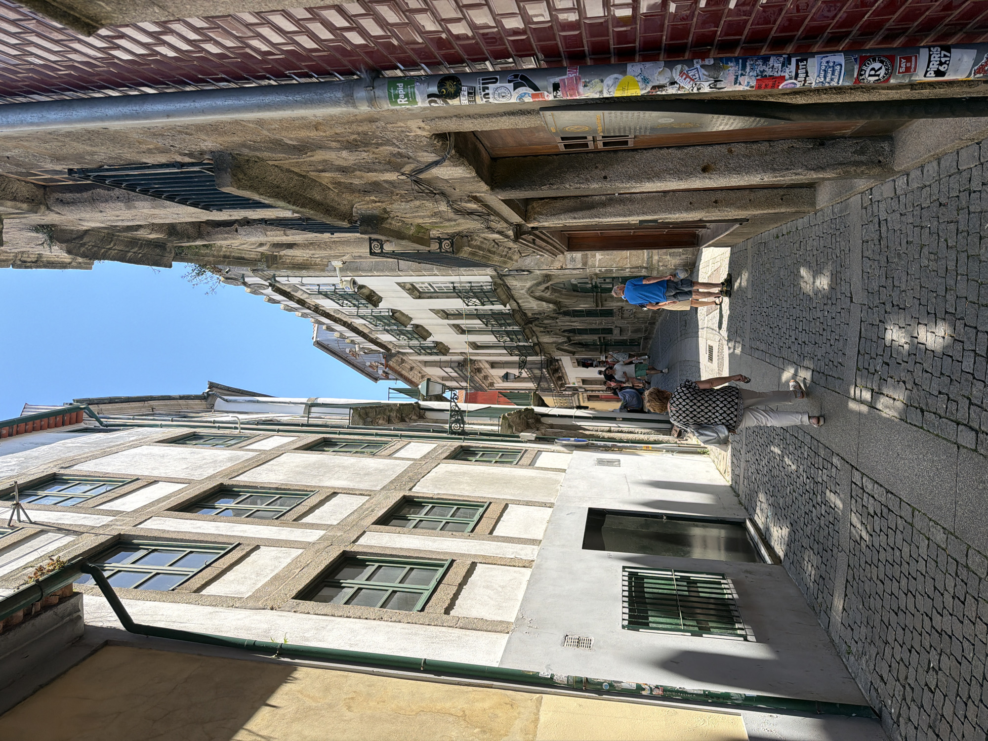 The image depicts a narrow, cobblestone street in Vila Nova de Gaia, Portugal, with buildings lining both sides. The sun is shining, casting distinct shadows on the ground. On the left, a building with a yellow facade transitions to a white and timber-framed structure with green-trimmed windows. A dark doorway and a barred window are visible on this side. On the right, a building constructed from rough stone features a metal pipe running vertically, covered in various stickers. This building has wooden doors and a red brick facade on the far right. In the middle ground, a woman wearing a patterned shirt and white pants walks away from the camera, carrying a light-colored bag. Further down the street, a man in a blue shirt and shorts is also walking away. Several other people are visible in the distance. The sky is clear and blue.