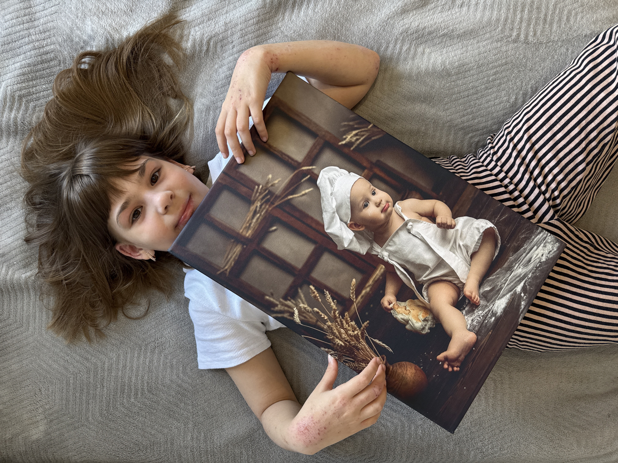 A girl lies on a gray textured surface, likely a bed or couch. She is holding a canvas print depicting a baby dressed as a baker. The girl wears a white shirt and black and pink striped pants. Her hair is brown and spread out on the gray surface. Her right arm is visible and has some red spots. The canvas print shows a baby wearing a chef's hat and an apron, surrounded by flour and holding a piece of bread. Wheat stalks and a pumpkin are also visible in the picture. The background of the print shows a window. The location is not apparent from the image and the city of Rusanovo, Russia, cannot be determined.