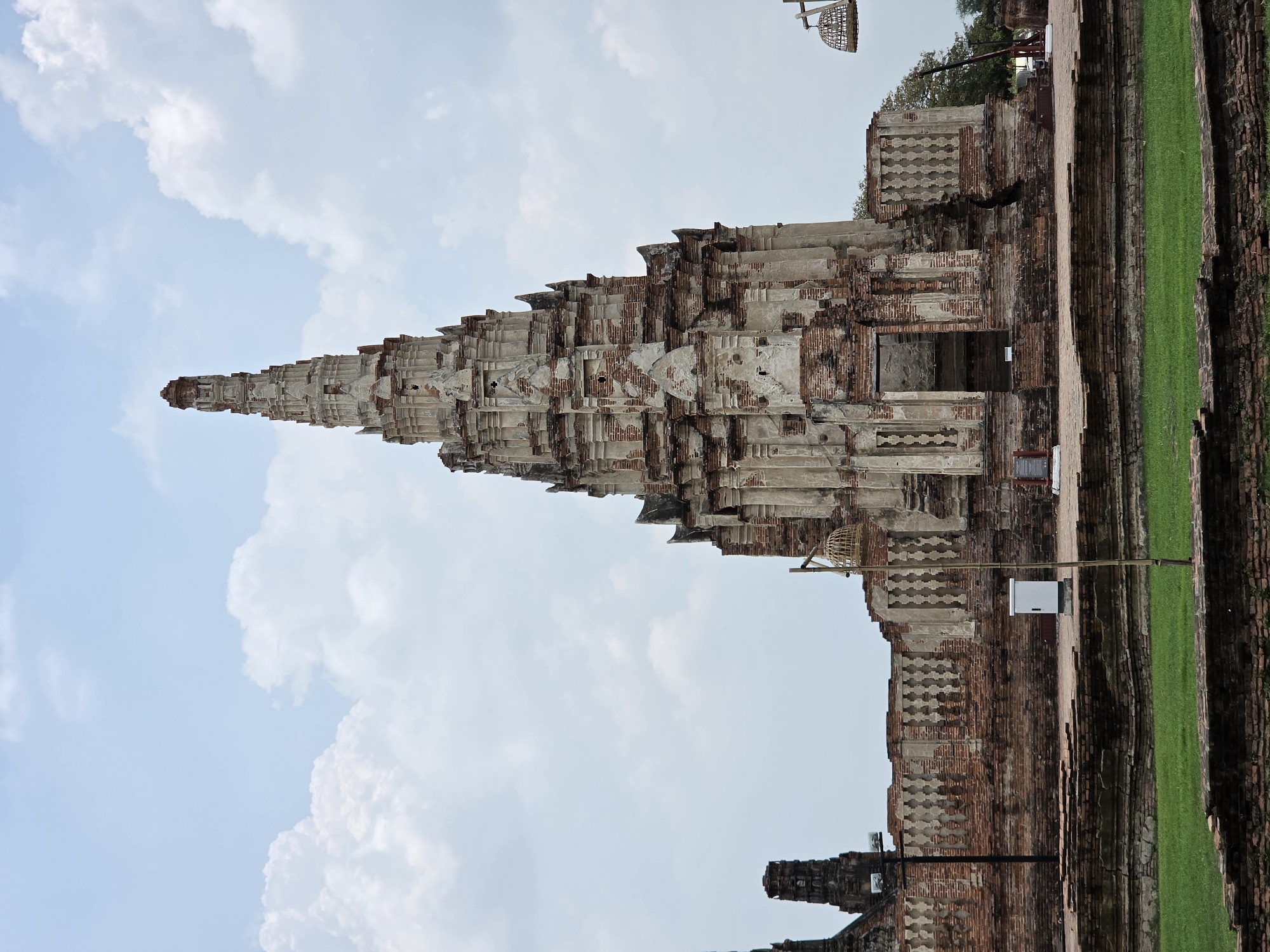 The image shows the temple ruins of Wat Chaiwatthanaram, located in the Ban Pom Subdistrict of Thailand. The central feature is a large prang, a tower-like structure, exhibiting a stepped, pyramidal form with detailed brickwork and weathered surfaces. A structure with a door is on the right side of the central prang. Adjacent to the prang, there are remnants of a brick wall and a strip of green grass. The sky is visible behind the prang with scattered clouds. The picture is framed vertically.