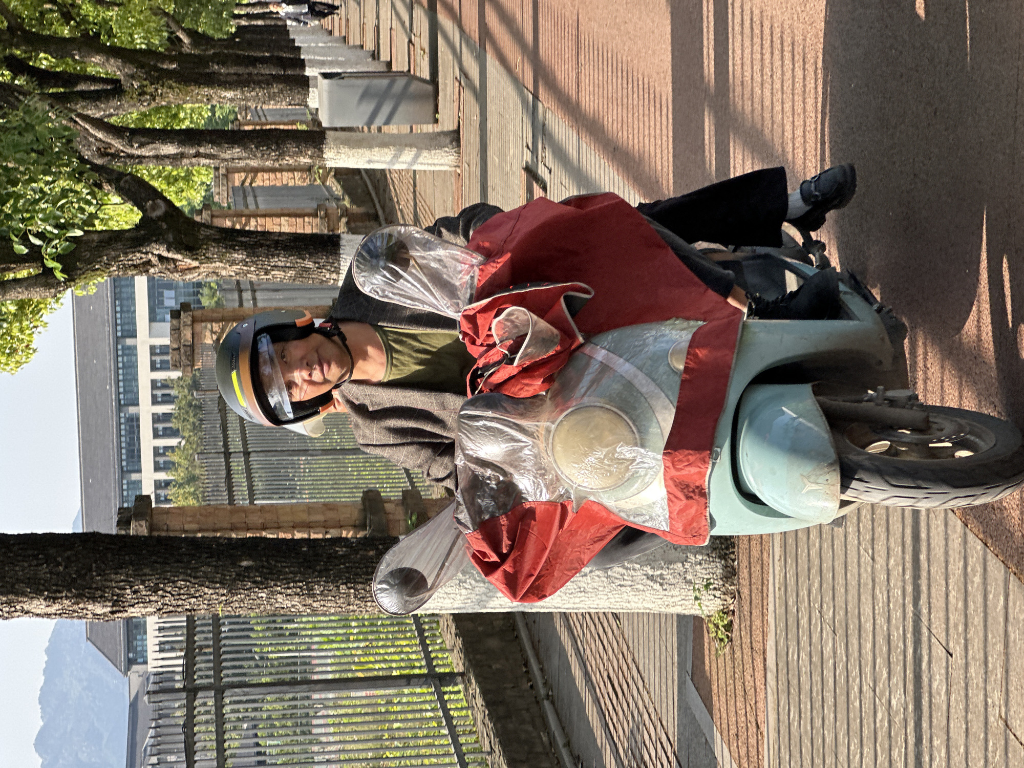An outdoor, eye-level photograph captures a man seated stationary on a light blue scooter on a paved path. The man, positioned centrally in the frame, wears a dark green t-shirt, a dark jacket, black pants, and a black helmet with a clear visor, looking directly at the viewer with a neutral expression. His hands rest on the handlebars, which are partially obscured by a large, red and clear plastic wind/rain shield draped over the front of the scooter.
The paved path beneath the scooter and extending into the background appears to be textured concrete or paving stones, marked by prominent, long shadows cast by trees and an unseen structure, indicating either early morning or late afternoon sunlight. To the left of the path, a row of mature trees with lush green foliage provides shade, their thick trunks visible along the edge. Behind the trees, a dark metal fence separates the path from several multi-story buildings, which are partially visible under a clear, bright sky. In the far background, faint outlines of mountains can be seen.
The scooter's front wheel is visible on the right, and the overall setting suggests an urban or peri-urban environment in sunny weather. The man appears to be taking a break or waiting. No discernible text is visible on any of the subjects or objects in the image. The location is Jiangjin, China.