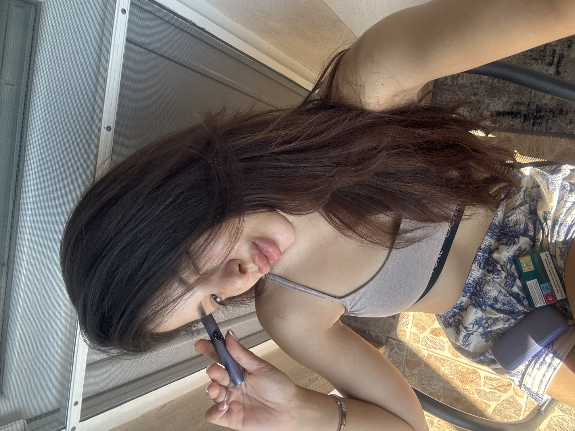 A young woman with long, dark, layered hair is taking a selfie outdoors. She is wearing a light grey sports bra and patterned shorts. She is holding a purple electronic smoking device to her temple with her left hand, and a pack of cigarettes is visible on her lap. The background appears to be a patio or balcony area with sunlight filtering through, suggesting an urban environment. The specific location in Plovdiv, Bulgaria, cannot be determined from the image alone.