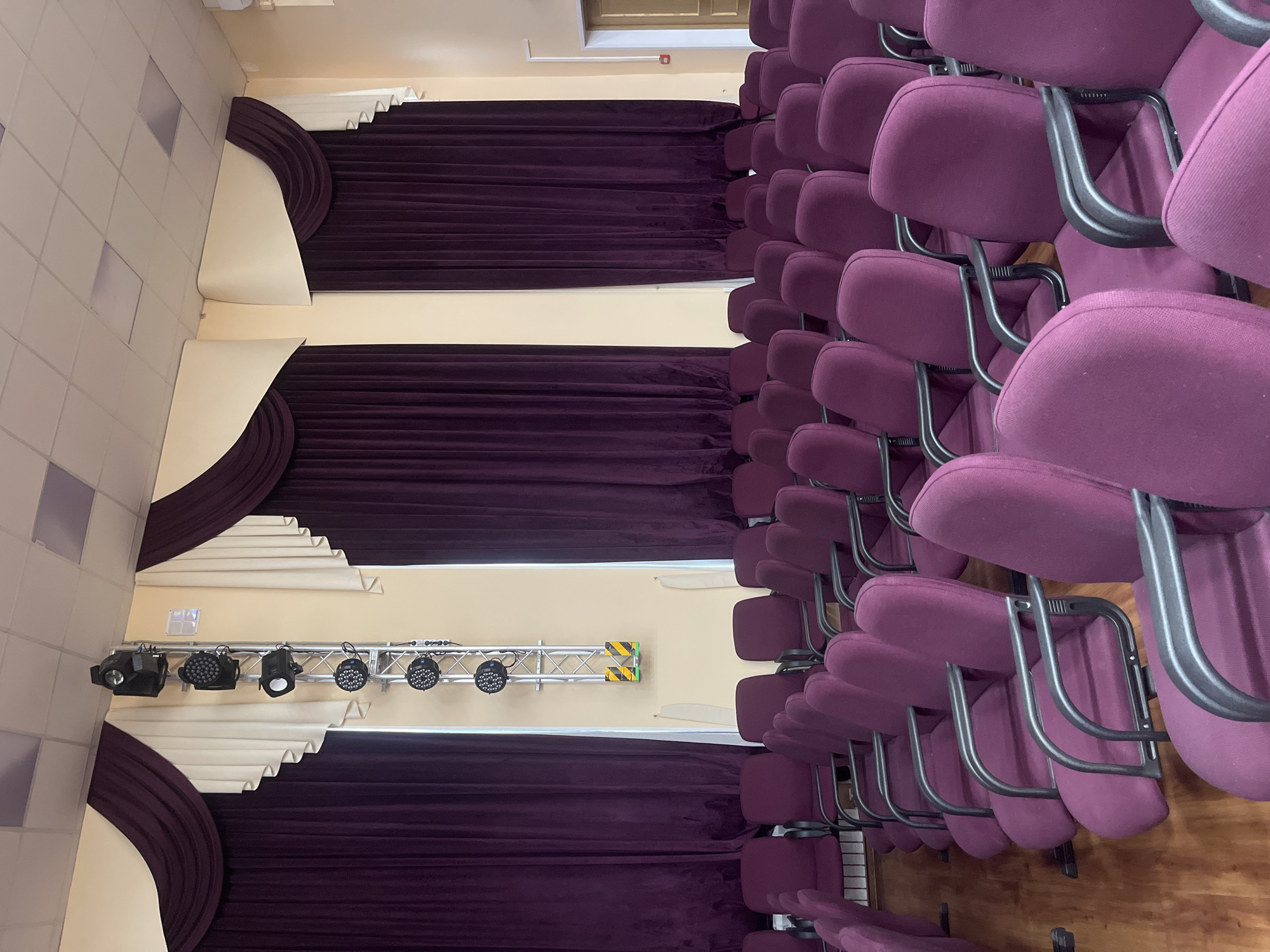 The image shows a room, likely a theater or auditorium, with rows of empty purple upholstered chairs. The chairs have black metal frames. The wall behind the chairs is decorated with alternating sections of purple curtains and white, pleated fabric. Stage lighting is mounted on a truss in front of one of the white fabric sections. The overall setting suggests a performance or presentation space. It's not possible to identify the specific location as Saint Petersburg, Russia, without further context or identifying features within the image.