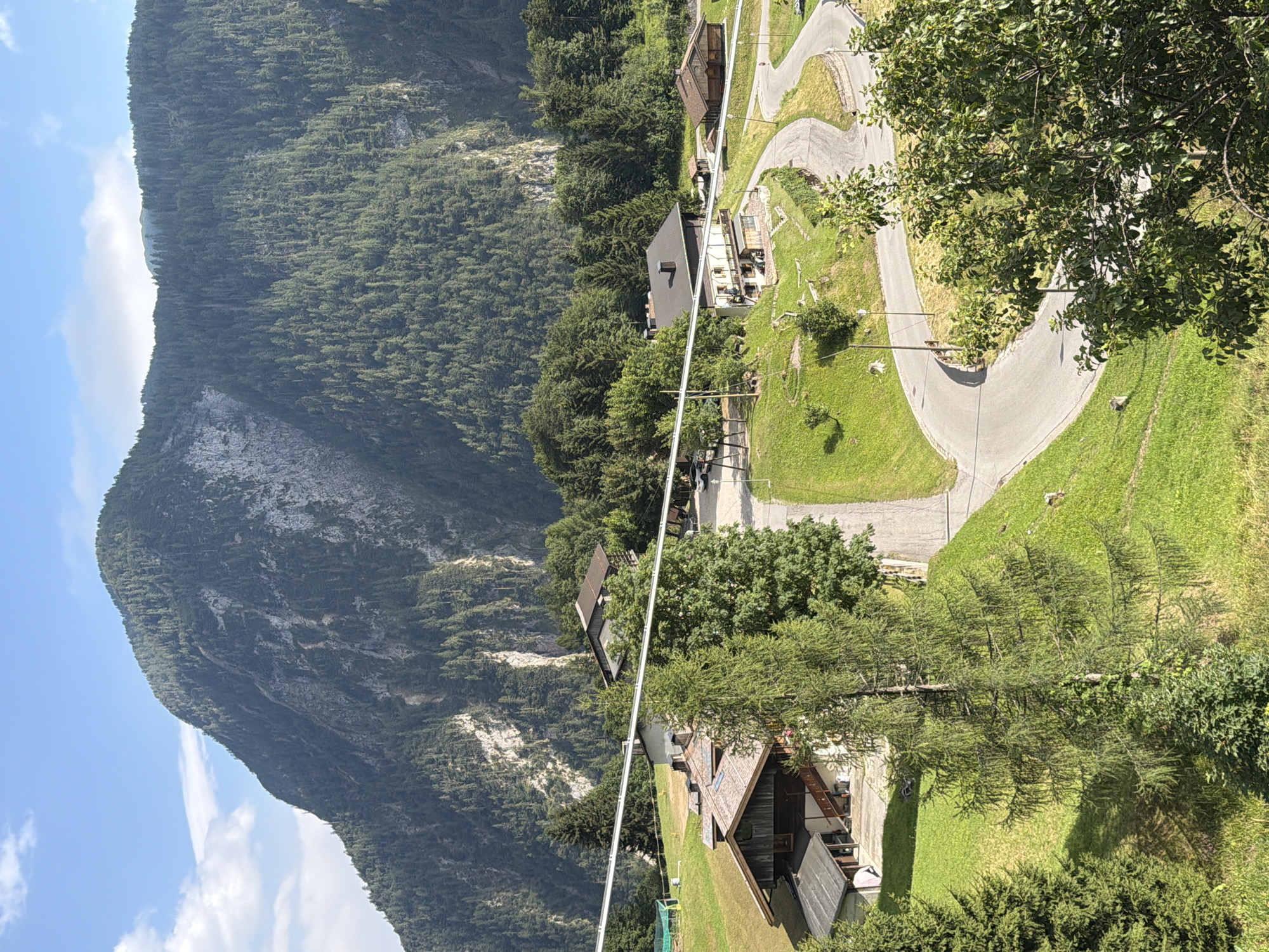 The image depicts a scenic mountain village in Sauris / Zahre, Italy. The foreground features a winding paved road curving through lush green grassy hills. Several traditional wooden houses with dark roofs are nestled into the landscape. In the mid-ground, more houses and trees are visible, with a large, imposing mountain covered in dense green forest rising majestically in the background. The sky is a clear blue with a few scattered white clouds. The overall impression is of a tranquil, idyllic mountain community bathed in sunlight.