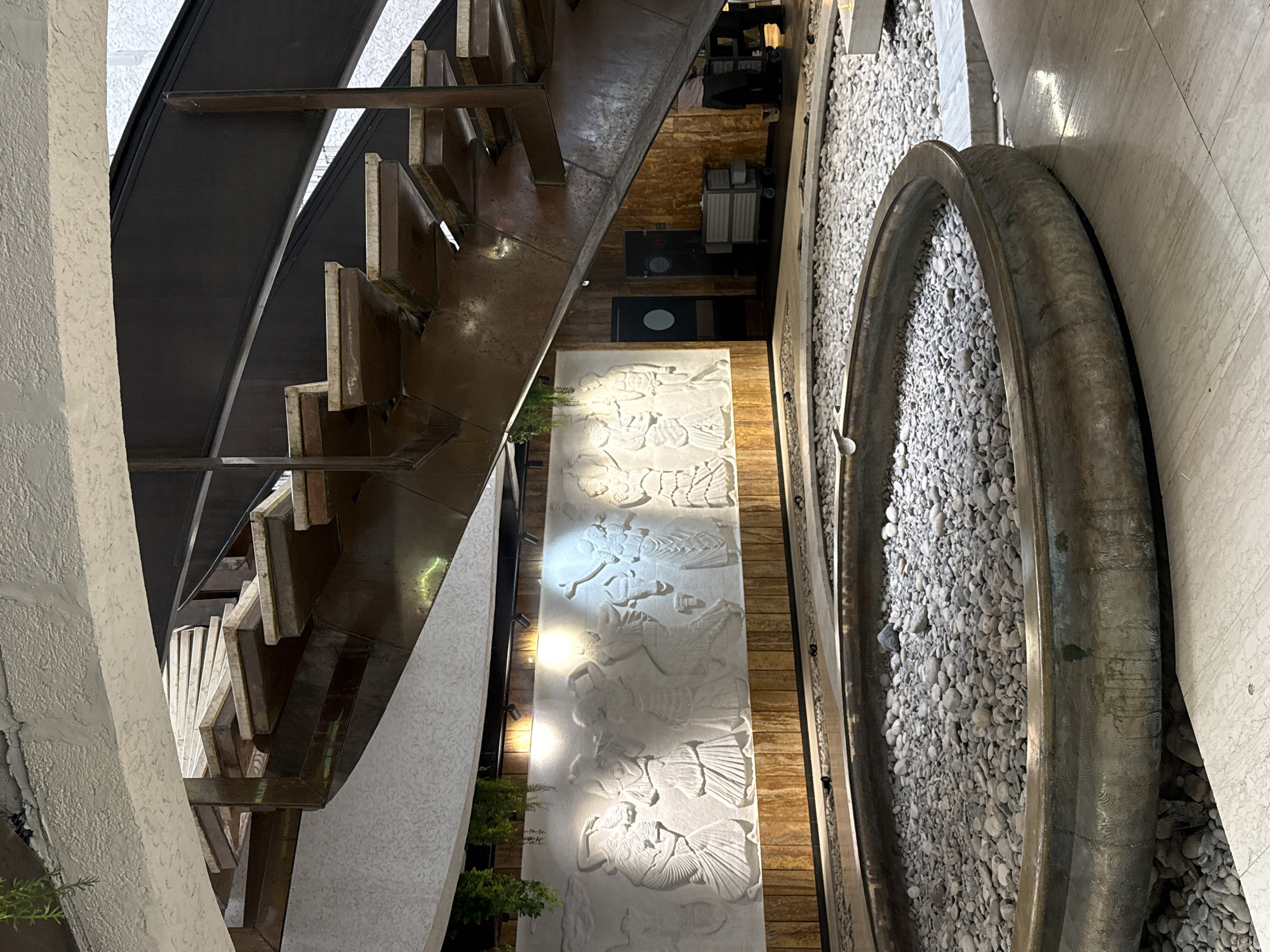The image depicts an indoor scene featuring a multi-level architectural design with a prominent spiral staircase. The staircase has wooden steps with a metallic, possibly copper or brass, frame and supports. Below the staircase, a large, circular basin filled with grey pebbles is visible, suggesting a decorative water feature or a dry landscaping element.
Adjacent to the pebble-filled basin and below the staircase is a wall adorned with a white bas-relief carving. This carving portrays several figures in a processional or celebratory scene, with detailed drapery and expressive poses. The wall is illuminated by spotlights, highlighting the texture and detail of the relief.
The environment appears to be a public space, possibly a hotel lobby, museum, or a restaurant, given the scale and decorative elements. The flooring in the foreground is polished stone or tile. In the background, behind the bas-relief, there is a wooden-paneled wall and doorways, with what appears to be service carts and staff members, indicating a functional area beyond the decorative space.
The image does not contain information to place it in the city of Yalta, Ukraine.