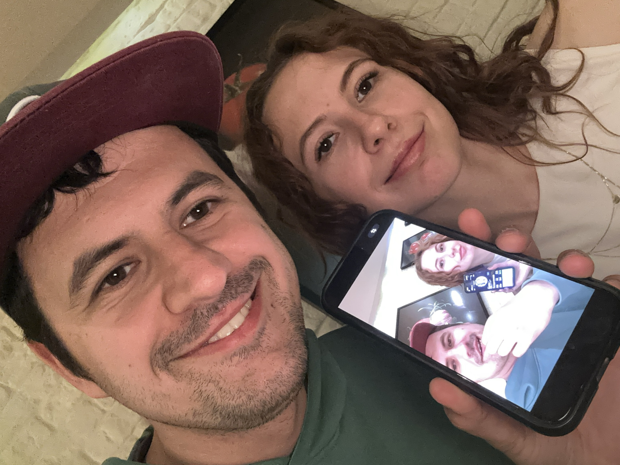 A selfie shows a man and a woman. The man is wearing a burgundy cap and a green shirt. The woman has curly red hair and is wearing a white shirt. The man is holding a phone displaying a previous selfie, in which he is holding a remote and the woman is holding another phone. The location is unknown and no information is present to place the location in the city of Podgorica, Montenegro.
