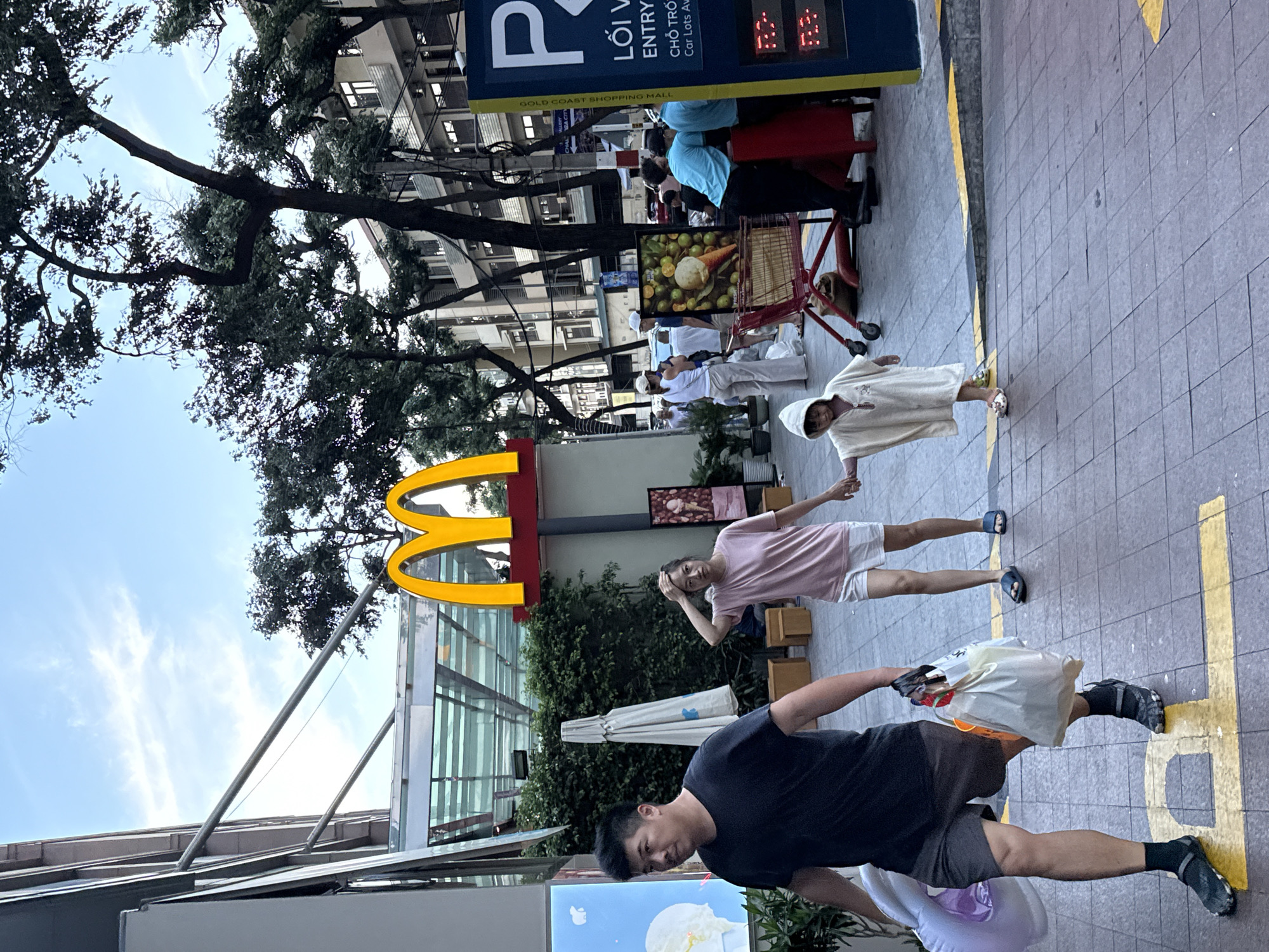 A group of people are walking on a paved sidewalk in front of a McDonald's restaurant in Nha Trang Ward, Vietnam. The McDonald's sign, featuring the iconic golden arches, is prominently displayed.
In the foreground, a man wearing a black t-shirt and shorts walks from left to right, carrying a large white inflatable ring and a plastic bag filled with groceries. He is wearing dark socks and sandals.
Behind him, a woman in a pink t-shirt and white shorts walks with her hand on her head. A young child, dressed in a white hooded garment, walks beside her, holding her hand.
In the background, other people are visible, some appearing to be shopping or waiting. A shopping cart with some items is present near a red payment booth. Several trees with dark foliage are scattered throughout the scene, and buildings with multiple windows line the street. The sky is cloudy. A tall blue and yellow sign with "P" and Vietnamese text is also visible on the right side of the image.