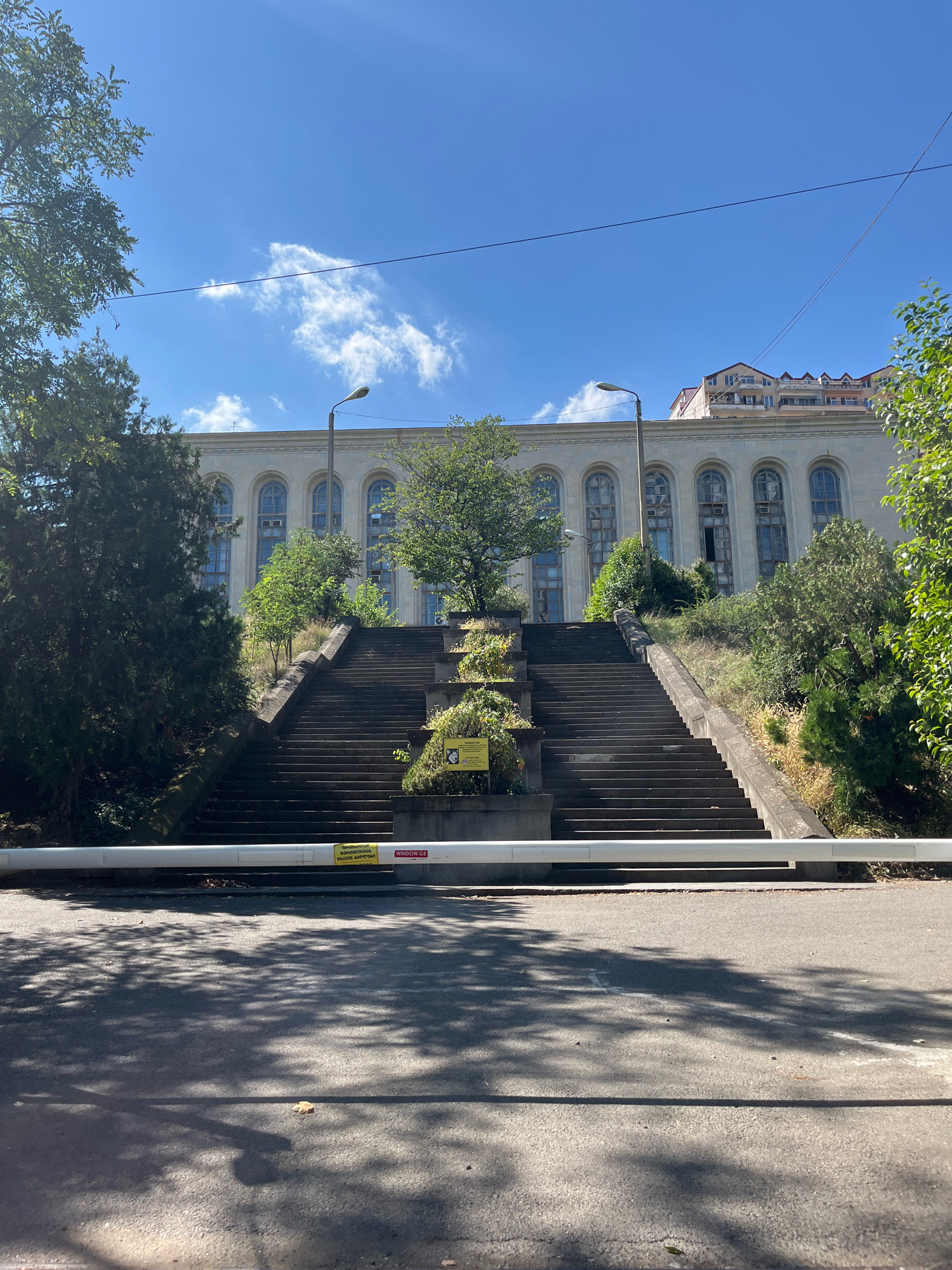 The image depicts an outdoor scene in Tbilisi, Georgia, featuring a grand, aged building with large arched windows. A wide, weathered stone staircase leads up to the building, flanked by overgrown vegetation and trees. At the base of the stairs, a white barrier gate is visible across a paved road, with shadows of tree branches dappled across the asphalt. The sky above is a clear blue with scattered clouds. The building itself appears to be of a classical architectural style, with ornate detailing around the windows and a long, continuous facade. The overall impression is one of an imposing, perhaps historically significant structure, showing signs of age and the passage of time.