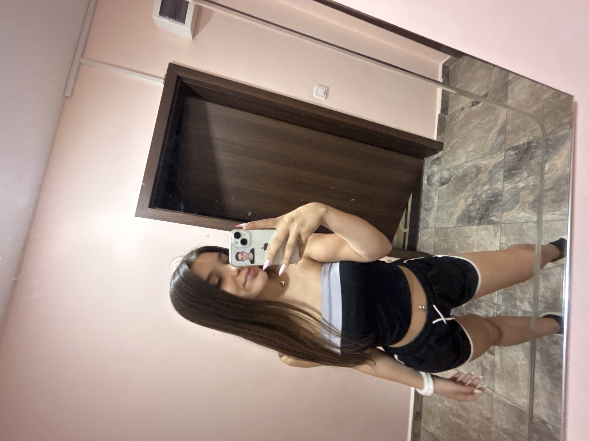 A young woman is taking a mirror selfie in what appears to be a hallway or room with a pink wall. She has long, dark hair and is wearing a black and white strapless top and black shorts with white trim. She is holding a smartphone in her right hand, and a small picture of a man is visible on the phone's case. A brown door is visible in the reflection of the mirror behind her. The floor is tiled with a gray and brown marble pattern. There is a light switch on the pink wall. The location is not specified as Shumen, Bulgaria.