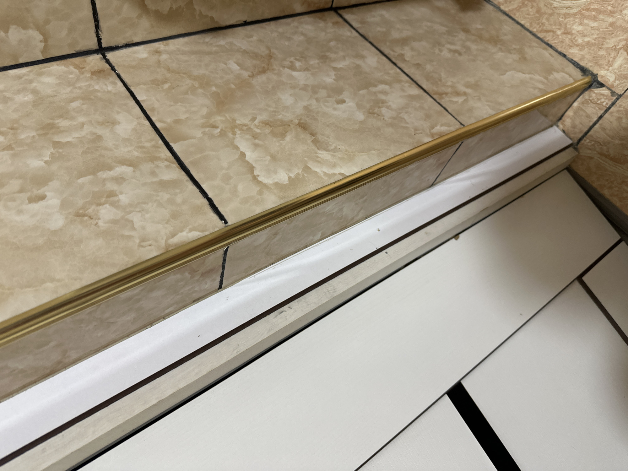 The image shows a close-up view of a corner where a white cabinet meets a tiled wall. A thin, gold-colored metallic trim runs vertically along the edge between the cabinet and the wall. The wall is covered in beige and brown marble-patterned tiles with black grout lines. The cabinet appears to be made of white, possibly lacquered, material with some visible seams. The lighting is somewhat dim, casting soft shadows. The scene depicts an interior corner, likely in a residential or commercial setting, such as a bathroom or a kitchen, within Fergana City, Uzbekistan. There are no people or animals visible in the image. The perspective is from a low angle, looking upwards into the corner.