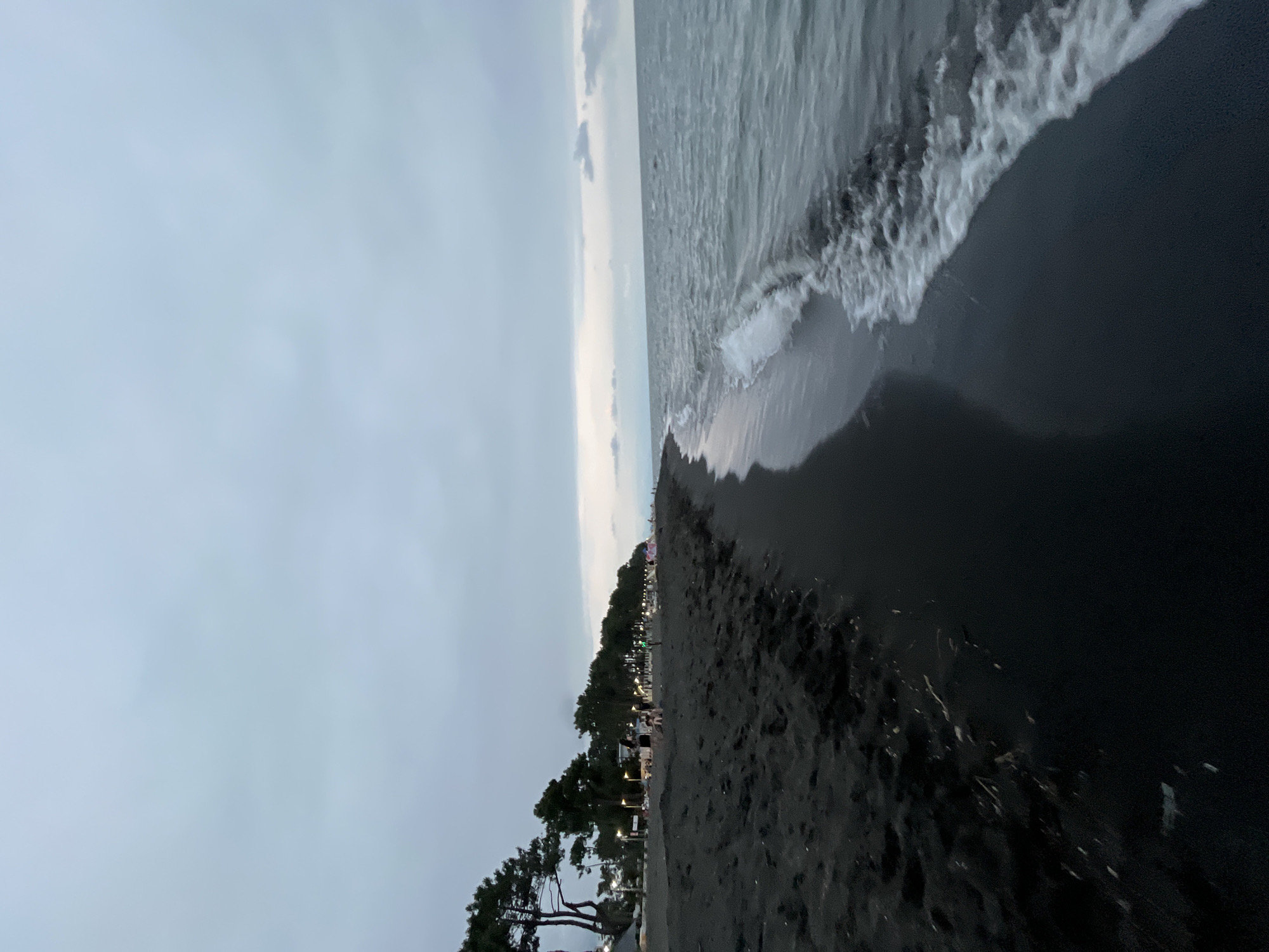 The image depicts a wide, dark sandy beach at dusk in Ureki, Georgia. The sky is overcast and a muted grey, with subtle hints of lighter clouds towards the horizon where the sea meets the sky. The sea is a calm, choppy expanse of grey-blue water, with gentle waves lapping at the shore, leaving white foam on the dark sand. A line of tall, slender pine trees borders the beach on the left, with some streetlights visible amongst them, indicating a developed area or promenade behind the beach. Several people are scattered along the edge of the beach and beneath the trees, some appearing to be sitting or walking. The overall impression is one of a peaceful evening at a coastal resort town.