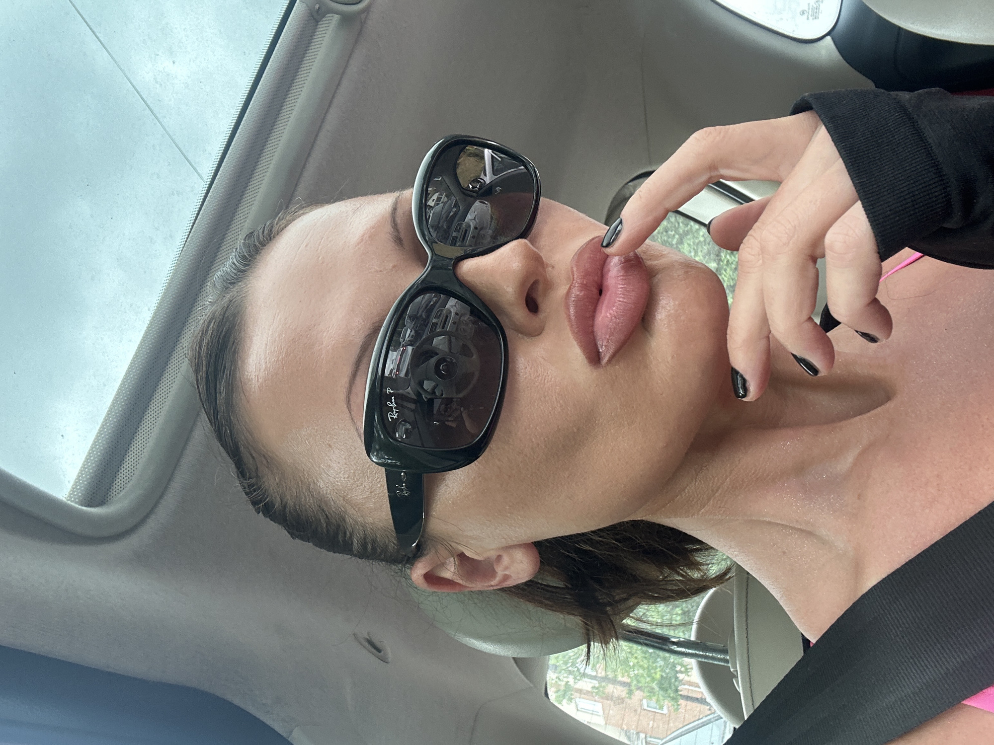 A woman wearing large black sunglasses and a pink top is in a car. She has her index finger touching her lips, which are painted with a glossy pink lipstick. Her nails are painted black. She has her hair pulled back. The interior of the car is visible, including a sunroof. The car is likely parked or moving slowly as buildings and trees are visible through the windows. There is no indication of the specific location within Tunbridge Wells, United Kingdom.