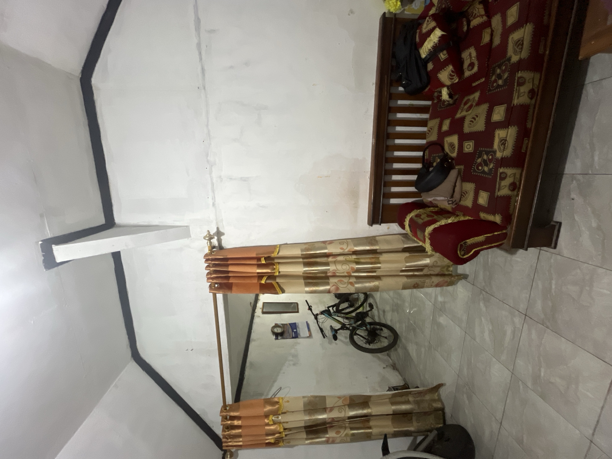 The image depicts the interior of a room, likely a living area, in a dwelling located in Pekalongan, Indonesia. The room features a sloped ceiling with exposed white beams painted black along the edges where they meet the walls. A wooden curtain rod stretches across the room, supporting two sets of light brown and gold striped curtains that frame an opening to an adjacent area.
Through the opening, a bicycle with a black frame and blue accents is visible, leaning against a white wall. On the wall, a calendar and a small framed picture are hung. The floor is tiled with light gray square tiles.
In the foreground, a wooden daybed with a dark wood frame is present. The daybed is covered with a patterned red and gold fabric. Two pillows, also in the red and gold pattern, rest on the daybed. A black backpack and a light brown bag are placed on the daybed, alongside the pillows. A portion of a red upholstered armrest is visible to the left of the daybed, and a glimpse of a yellow object, possibly a flower, is seen to the right. The walls of the main room are painted white and appear to have some water stains or discoloration.
