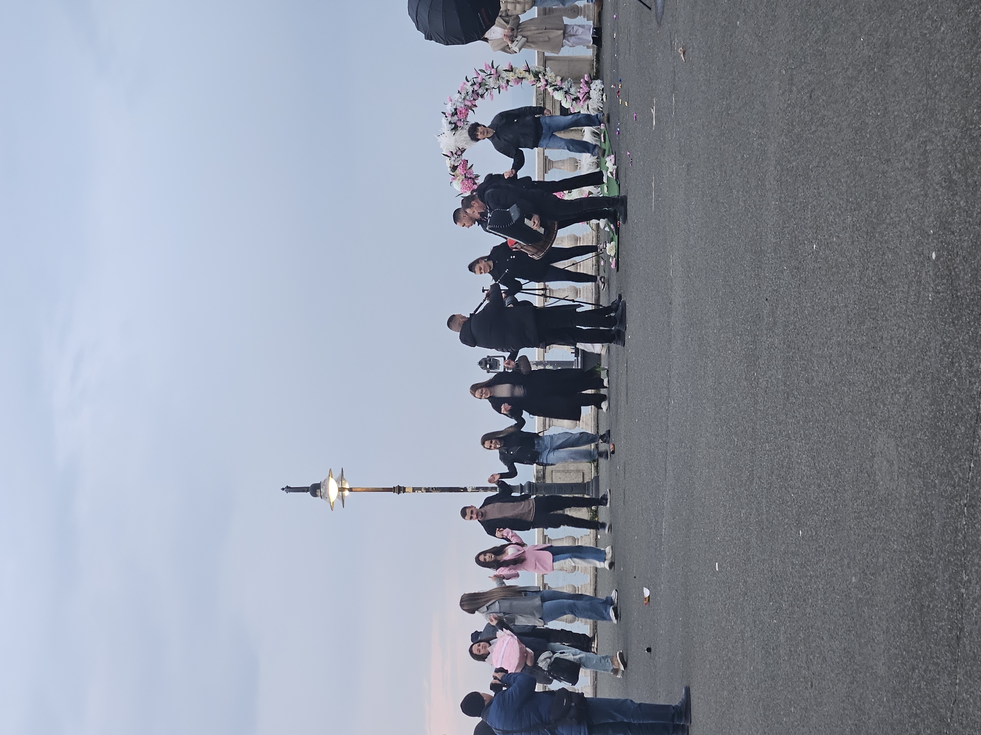 The image depicts a group of people standing on a paved surface next to a body of water, possibly the Black Sea. The location is Burgas, Bulgaria. The people are lined up, seemingly posing for a photo in front of a floral arch. Some individuals appear to be musicians, holding instruments such as an accordion and what seems like a clarinet. The image also shows a light post along the side. The sky is overcast, creating soft light.