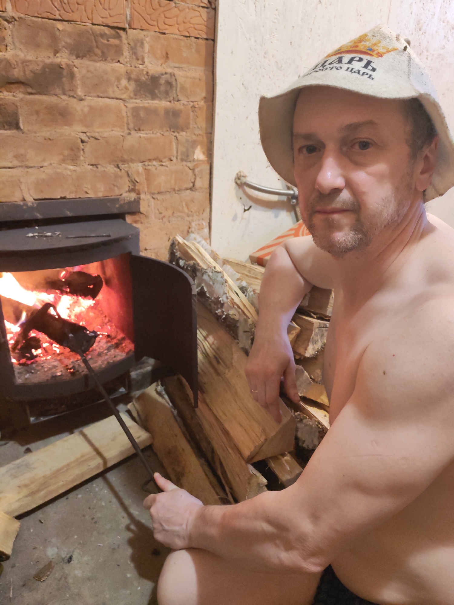 A man is sitting next to a wood-burning stove in what appears to be a sauna. He is shirtless and wearing a hat with Russian text on it. The stove door is open and burning wood is visible inside. There is a poker tool in his hand, and he is stirring the fire. A pile of firewood is next to him. The walls are brick and painted white.