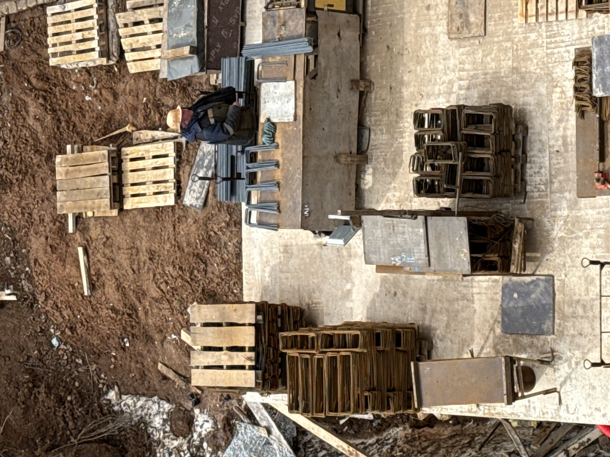 This aerial, high-angle shot captures an outdoor construction or material staging area. The ground is split between a dark brown, uneven, muddy earth surface on the left and a lighter, smoother, concrete or compacted gravel area on the right.
One individual, wearing a light-colored hard hat, a dark jacket, and light-colored pants, is visible in the upper middle of the frame. They are crouched or sitting on the lighter ground, appearing to be working with or organizing the materials around them.
The site is filled with various construction components. Numerous wooden pallets, some stacked, are scattered across both the muddy and lighter ground sections. A large quantity of steel reinforcing bars (rebar) is present; many are pre-bent into U-shapes or rectangular stirrups, neatly stacked in piles on the lighter surface. Straight rebar pieces are also visible. Large, rectangular panels, likely formwork made of wood or metal, are positioned around the working area. On one panel, the text "PLY" and parts of "SYD" are visible. Smaller metal plates and other miscellaneous tools or debris are also scattered throughout.
The scene is brightly lit, suggesting daytime, though the absence of prominent shadows could indicate an overcast sky or a high sun angle. The juxtaposition of raw earth and organized construction materials points to an active building site.