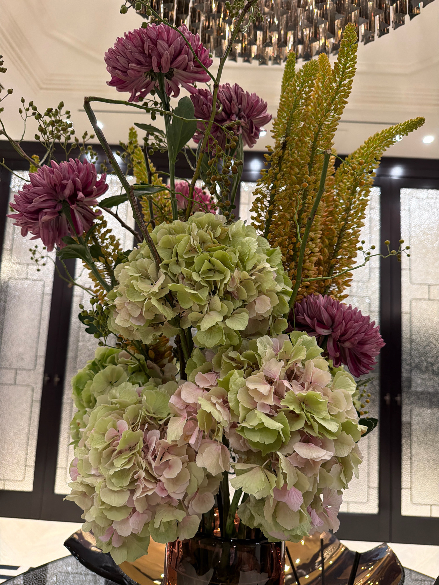 This is a close-up, vertical, indoor photograph of a floral arrangement. The arrangement features large, pale green and pink hydrangeas in the foreground, interspersed with spiky, golden-yellow flowers and deep purple, chrysanthemum-like blooms. Small, green berries on thin stems are also visible, adding a delicate texture. The flowers are displayed in a dark, possibly glass or ceramic, vase which is partially visible at the bottom of the frame. The background is out of focus, showing a dimly lit room with what appears to be a large window or glass partition with a grid pattern and a modern, multi-tiered chandelier overhead. The lighting suggests an interior setting, possibly a hotel lobby, a formal dining room, or a high-end retail space. There are no people or animals present in the image. The overall impression is one of elegant and sophisticated floral decor. There is no information in the image to indicate the location is Moscow, Russia.