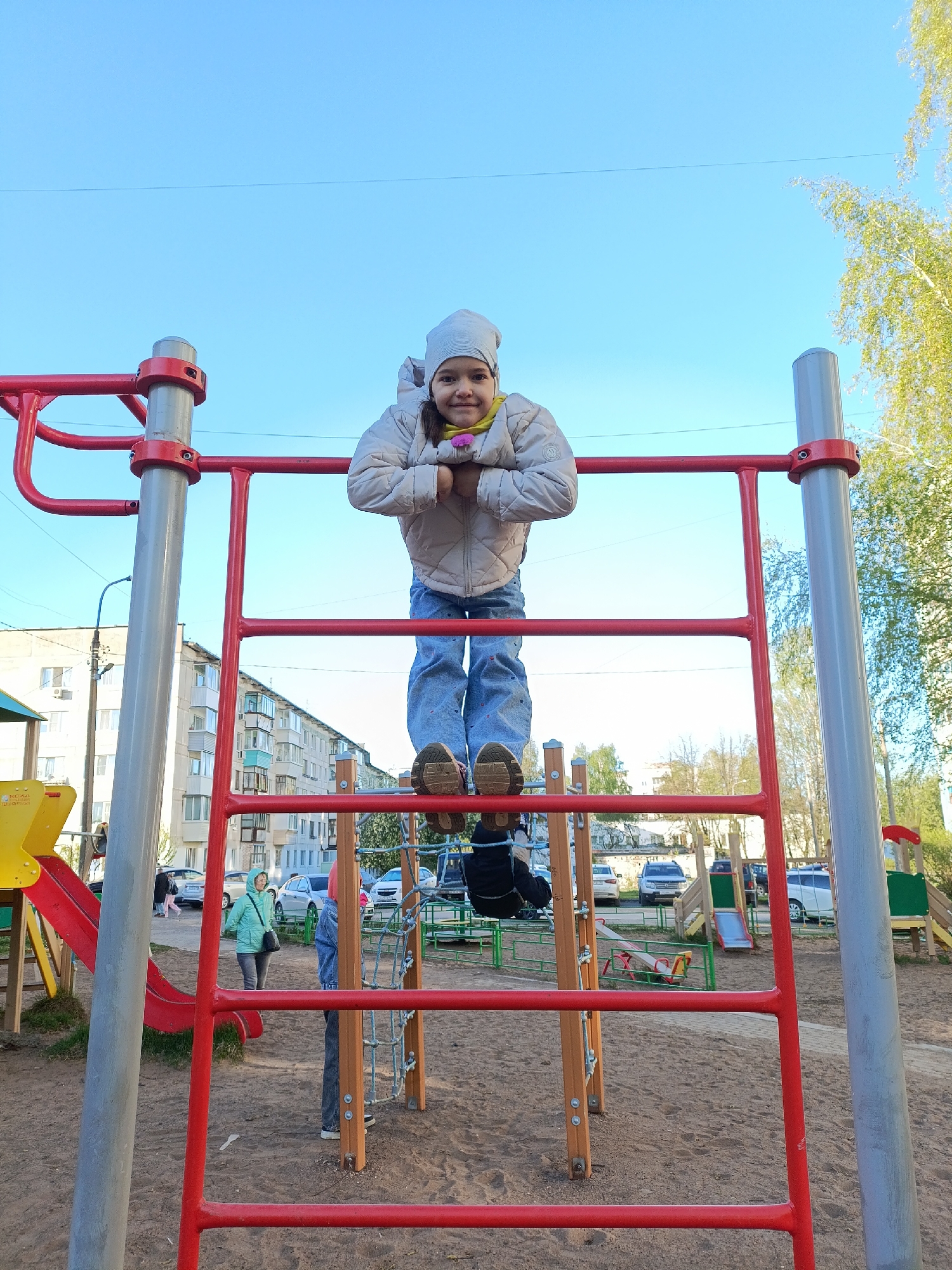 A girl in a light jacket, blue jeans, and a gray hat stands on a red metal ladder structure in a playground. Her arms are resting on the top bar of the ladder. The playground has a sand surface. Other playground equipment, including a slide, are visible in the background. Beyond the playground, there are apartment buildings, indicating an urban environment, specifically the city of Chaikovsky, Russia. Several people are walking in the background. Parked cars are present near the buildings. The sky is blue.