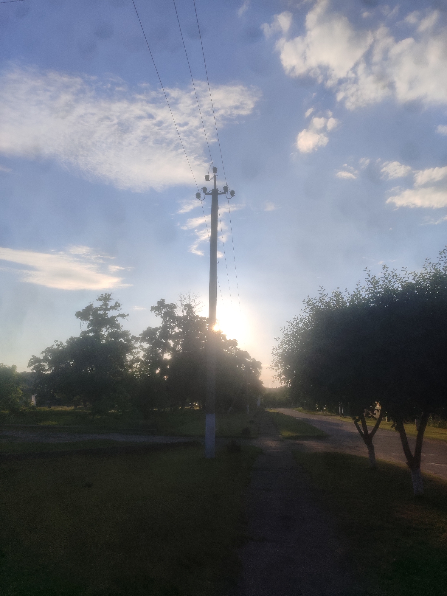 The image depicts a rural or suburban landscape with a prominent utility pole and overhead power lines stretching across a bright, partly cloudy sky. The sun is low in the sky, causing lens flare and a luminous glow behind silhouetted trees. A paved path or road is visible in the foreground, bordered by grassy areas and more trees. The overall scene suggests a natural environment with man-made infrastructure, possibly at dusk or dawn due to the lighting.