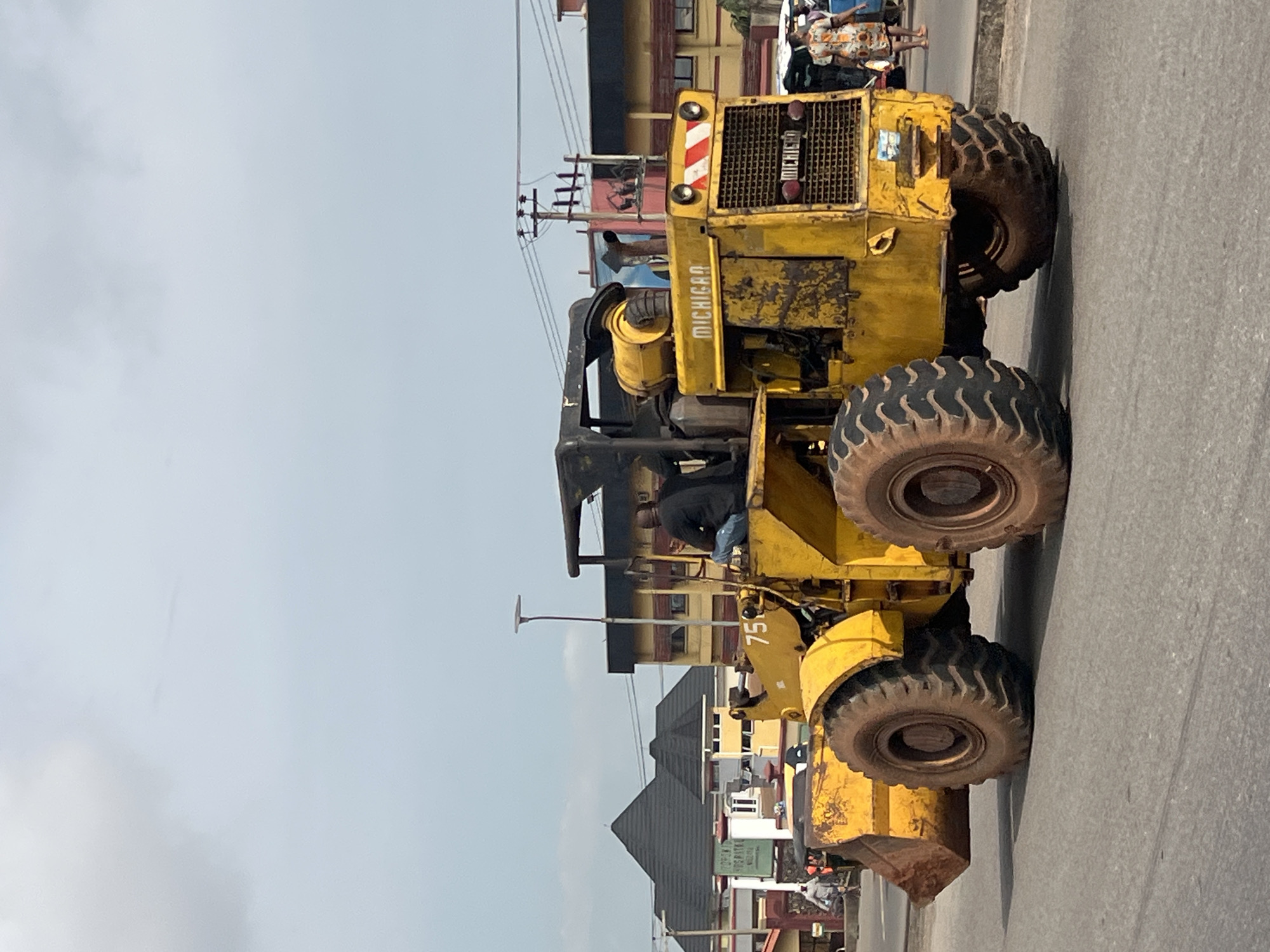 In Onitsha, Nigeria, a yellow Michigan front-end loader is parked on a paved road. A man is seated in the driver's seat of the loader. The loader has a large bucket attached to the front and four large, rugged tires. In the background, there are buildings with distinctive peaked roofs and a variety of signs, including one that appears to read "MICHIGAN." Power lines and utility poles are visible. A woman in a patterned dress is walking on the sidewalk. The sky is overcast.
