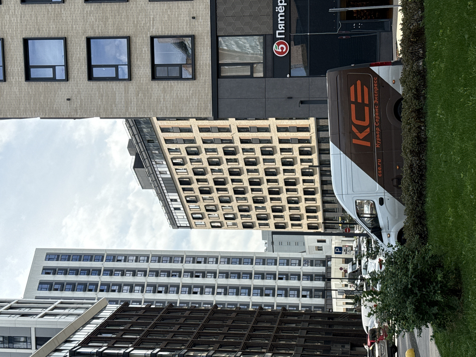 This is a street-level, eye-level medium shot of a street scene in Moscow, Russia, on a partly cloudy day. In the foreground, a white and brown delivery van with the logo "KCE Курьер Сервис Экспресс" is parked on the right side of the frame, partially obscured by a bush and green grass. The van has the website "cse.ru" printed on its side. To the left of the van, a row of parked cars and a street sign are visible.
The background features several modern high-rise buildings. On the right, a beige brick building with a dark facade on the lower level displays a red circular logo with the number "5" and the text "Пятёрка" (Pyaterochka, a Russian supermarket chain) to its right. Further left, a light-colored building with a grid of numerous windows is prominent. The leftmost buildings are darker, with a more complex facade design and many windows. The sky is overcast with scattered clouds. The overall impression is of a developed urban environment with a mix of residential and commercial buildings.