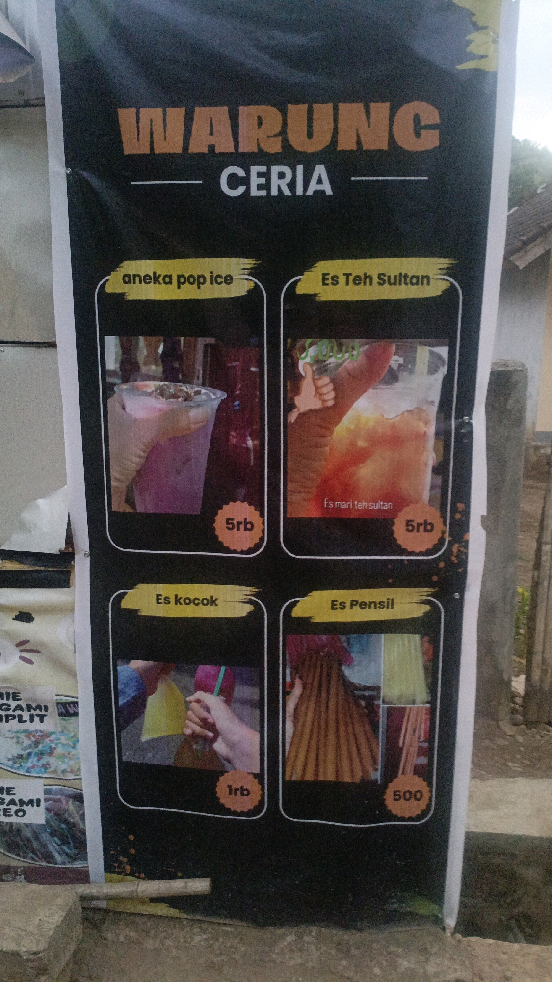 The image is a photograph of a banner advertising a food and beverage stall called "WARUNG CERIA" in Taliwang, Indonesia. The banner is black with orange and white text and features four product offerings with accompanying images and prices.
The top section of the banner displays "WARUNG" in large orange letters and "CERIA" in white letters below it, with a thin orange line separating them.
The first product, "aneka pop ice," is shown in a plastic cup filled with a pink beverage topped with sprinkles. The price is indicated as "5rb" in a rounded orange tag.
The second product is "Es Teh Sultan," which is a refreshing iced tea drink in a plastic cup with ice, and a hand giving a thumbs up next to it. The text "Es mari teh sultan" is visible at the bottom of the image. The price is also "5rb".
The third product is "Es kocok," which appears to be ice drinks in plastic packaging with straws, held by a person's hands. The price for this item is "1rb".
The fourth product is "Es Pensil," showcasing various colorful ice popsicles, some of which are long and thin. The price is listed as "500".
The banner is hung outdoors, likely on a street or sidewalk, as suggested by the concrete ground and a portion of a wooden post visible on the right side of the image. There is also a glimpse of foliage in the background. The overall impression is that of a casual, local business catering to the community.