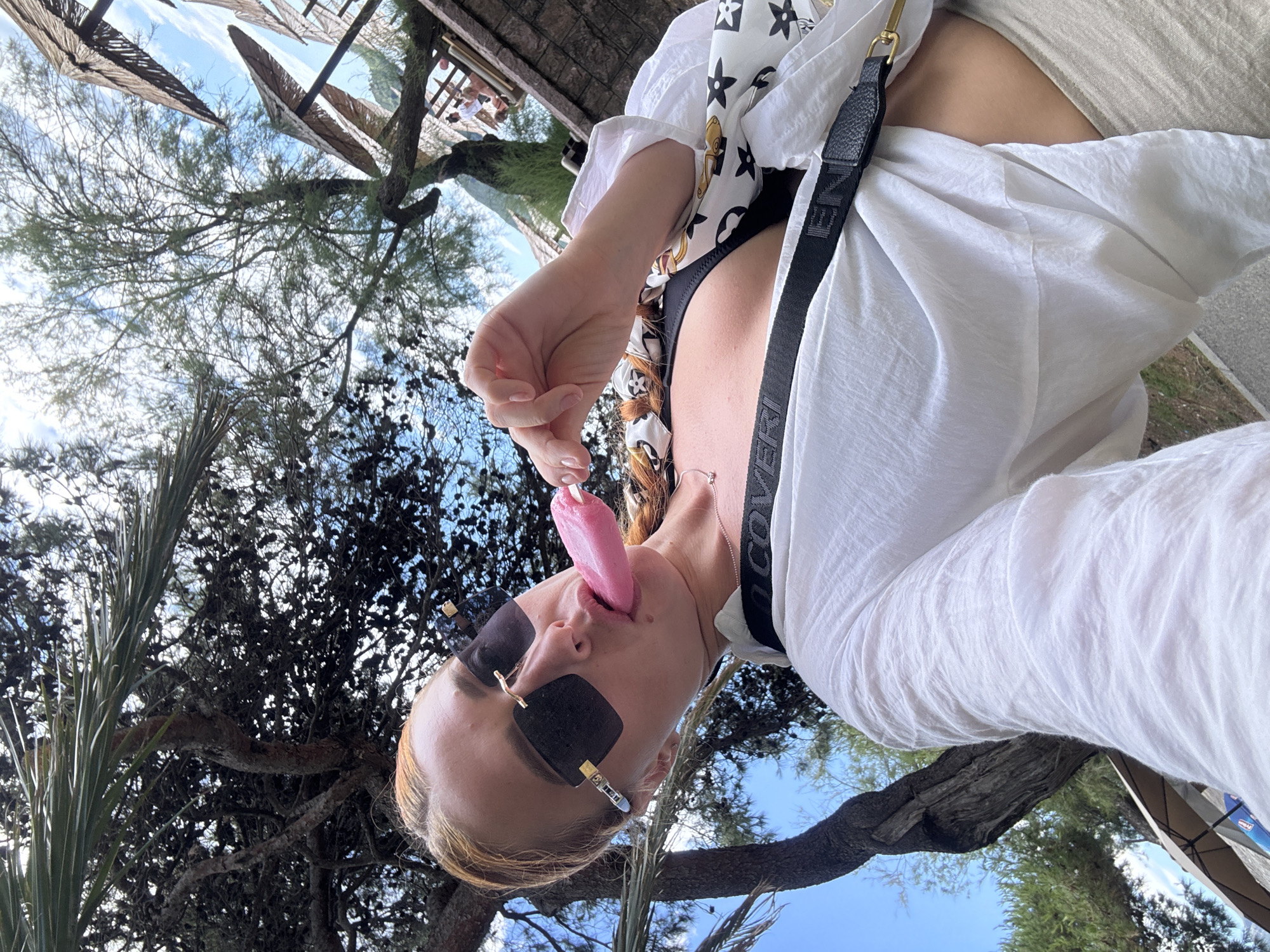 An individual with blonde hair, wearing sunglasses and a white open shirt over a black bikini top, is holding a pink popsicle to their tongue. The scene appears to be outdoors, with trees, foliage, and umbrellas suggesting a beach or resort setting, possibly in Budva, Montenegro, given the context. The background shows a blue sky with scattered clouds and hints of distant structures and greenery. A black strap with embossed lettering is visible across the individual's chest.