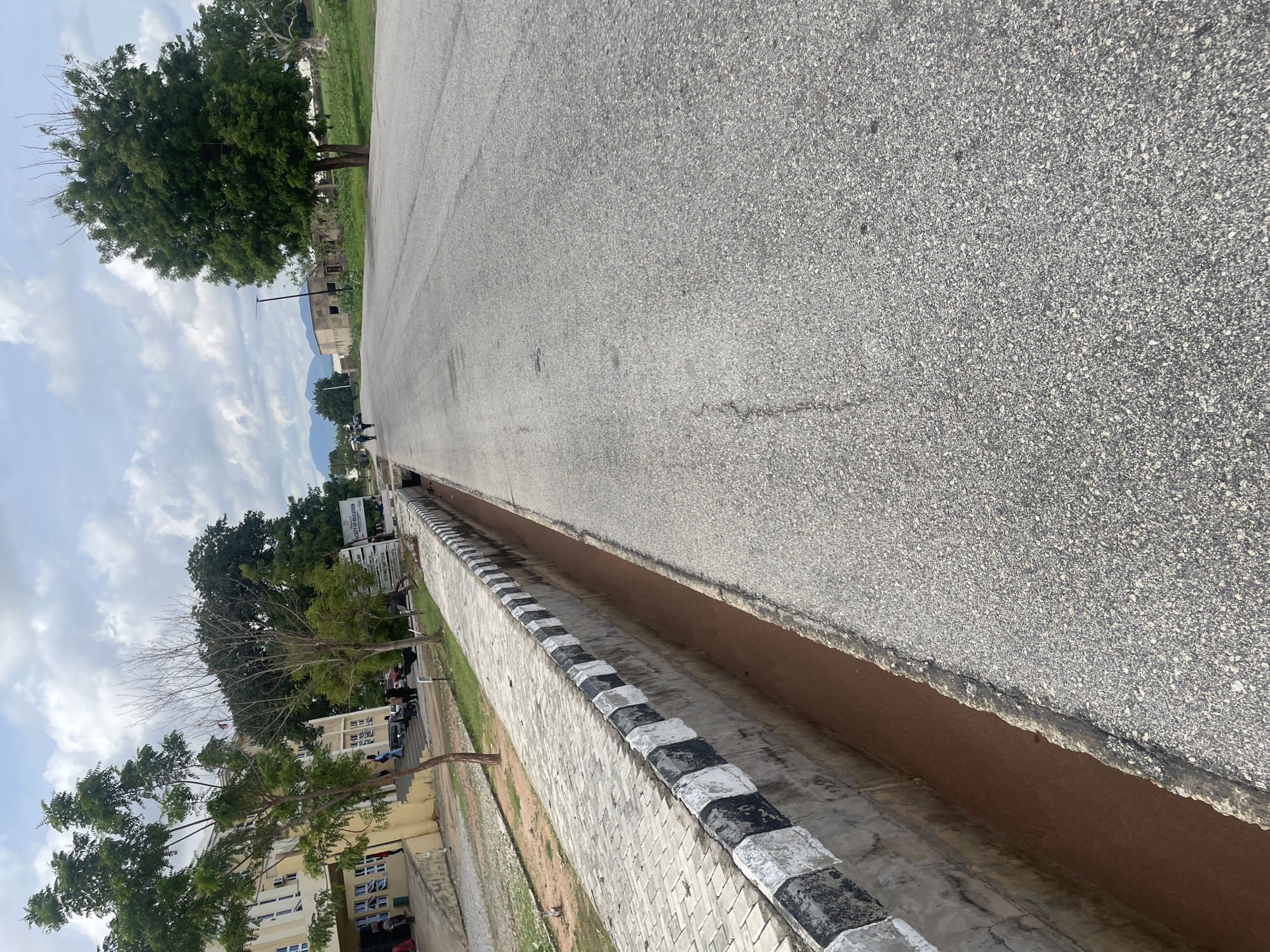 An overcast day frames a road in Jalingo, Nigeria, flanked by a concrete drainage ditch and a sidewalk. The road, a light grey asphalt surface, displays visible cracks and wear. On the left, a building with a pale yellow facade and numerous windows stands behind a row of trees. A sidewalk constructed of interlocking grey bricks runs parallel to the road, separated by a concrete wall adorned with alternating black and white painted blocks. The drainage ditch is filled with reddish-brown earth. In the distance, more buildings and trees are visible, with mountains forming a backdrop under the cloudy sky. A few individuals can be seen walking and standing near the building and on the sidewalk, and a truck is parked on the side of the road.