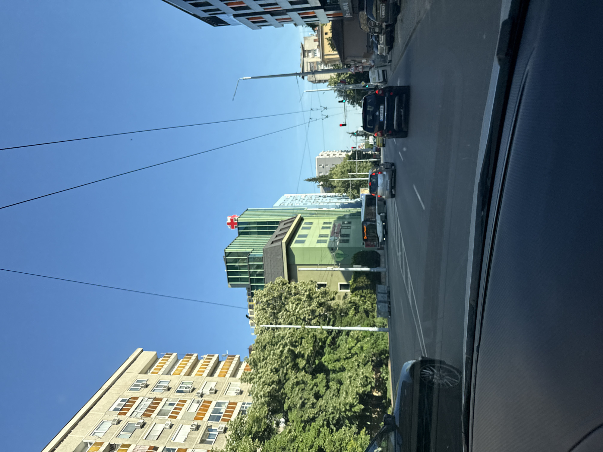 The image shows a street scene in Stara Zagora, Bulgaria, viewed from inside a vehicle. The sky is clear and blue. On the left, a tall, beige apartment building with orange-framed balconies and numerous windows is visible. Adjacent to it is a dense canopy of green trees. Further down the street, a green building with several windows is present, and behind it, a taller, modern building with a green facade and a red cross symbol on top. The street is paved and has multiple lanes. Several cars are on the road, including a black SUV in the foreground. A bus is also visible in the distance. On the right side of the street, there are parked cars and buildings. Traffic lights are visible, indicating a junction. The overall impression is a daytime urban environment in Bulgaria.