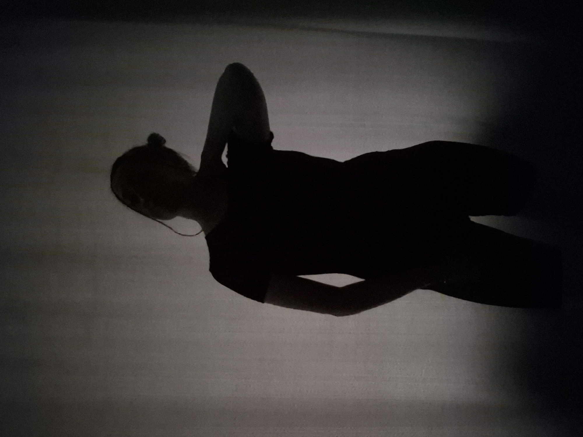A silhouetted figure lies on a light-colored surface, possibly a floor, in a darkened room. The person appears to be wearing clothing, with a bun hairstyle visible. One arm is raised and bent at the elbow. The environment is dimly lit, with strong contrast between the light-colored surface and the dark silhouette of the person. The location in Chaikovsky, Russia, cannot be determined from the image.