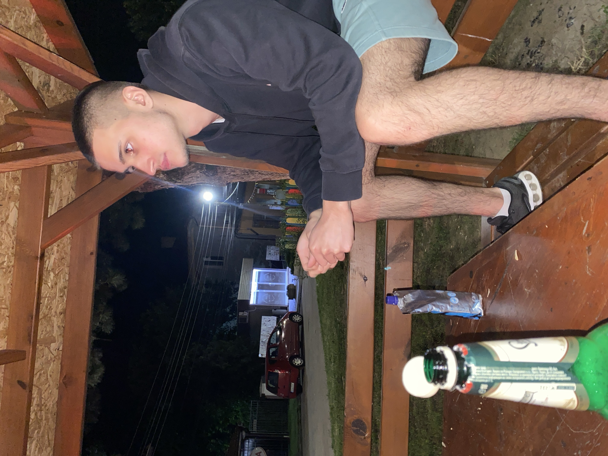The image depicts a young man sitting on a wooden bench in what appears to be an outdoor seating area with a wooden roof structure. He is wearing a black hoodie and light blue shorts. His legs are bare and hairy. In the foreground, on a wooden table, there is a green bottle with a white label, a white cap on the bottle opening, and a silver pouch. Behind the bench, there is a grassy area, a street with a red car parked, and a building with illuminated windows. The scene appears to be at night, indicated by the dark sky and streetlights. The location is identified as Yasen, Bulgaria.