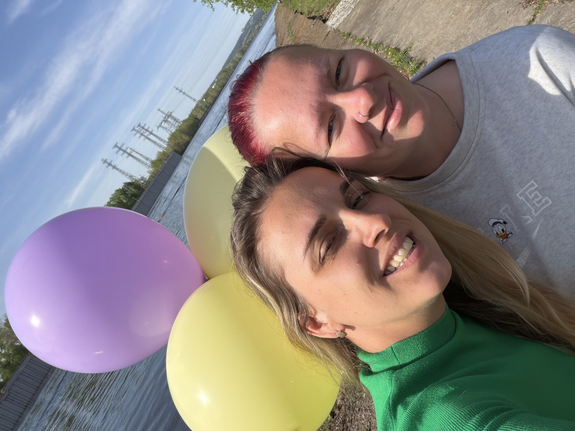 Two women are posing for a selfie outdoors, holding a purple and a yellow balloon. The woman in the foreground is wearing a green turtleneck shirt and has blonde hair. The woman behind her has short, red hair and is wearing a light gray sweatshirt with a cartoon character on it. In the background, there is a body of water with power lines and trees visible. The location is the city of Chaikovsky, Russia.