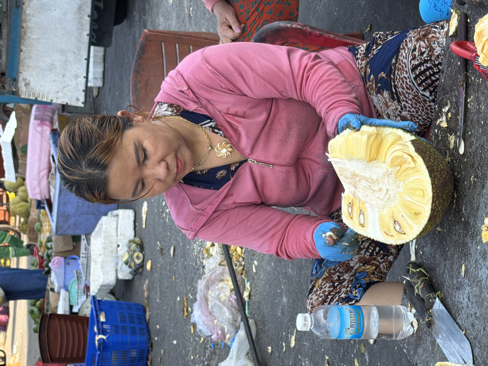 A woman is sitting on a red chair outdoors, preparing a large jackfruit. She is wearing a pink hooded jacket, a floral patterned top, and patterned pants. She has blue gloves on and is using her hands to pull apart the jackfruit. A bottle of Sanna brand water and a knife are on the ground in front of her. The scene appears to be a market setting with various crates and fruits visible in the background. This outdoor market likely takes place in the city of Nha Trang, Vietnam, known for its vibrant street markets.