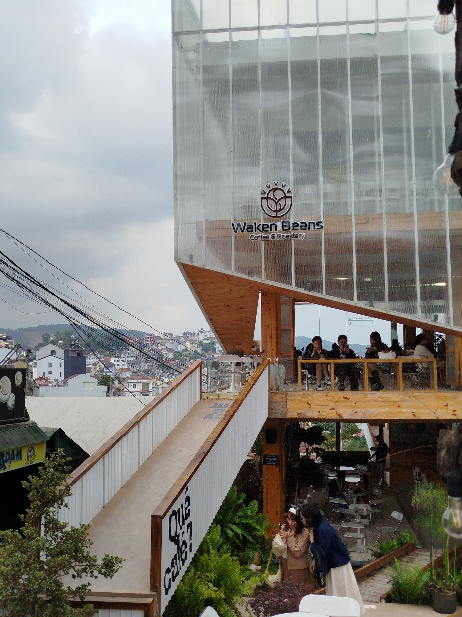 The image displays a multi-level modern coffee shop, "Waken Beans Coffee & Roastery," situated on a hillside, overlooking a dense urban landscape, characteristic of Da Lat, Vietnam.
The main building features a distinctive contemporary design. The upper section is encased in a translucent, possibly perforated, material or glass, giving it a bright, airy aesthetic. This section cantilevers over a lower, open-air or semi-enclosed area constructed primarily from exposed wooden beams and planks, creating warm, natural tones. An angled, elevated concrete walkway with white corrugated metal and wooden railings provides access, winding upwards towards the main structure.
Numerous people are visible enjoying the cafe. On an outdoor terrace of the upper level, a group of five individuals are seated at white tables, mostly engaged with their mobile phones or in conversation. Another person is partially visible further back, potentially a staff member. On a lower, more garden-like level, two women stand together, one holding a phone, while other patrons are seated at scattered tables.
The background reveals a bustling townscape with numerous houses and buildings clustered across rolling hills, extending into the distance. A dense array of black power lines crisscrosses the mid-ground sky, leading towards the residential areas. The sky is overcast with soft, gray clouds, indicating a cloudy day, rather than bright sunshine.
Notable details include lush green foliage and ferns integrated into the landscaping around the wooden structures, adding a natural touch. Bare filament light bulbs hang from various wooden supports. An open umbrella is visible on the upper terrace.
Visible text on the main facade clearly states "Waken Beans Coffee & Roastery" accompanied by a stylized bean or leaf logo. On the side of the elevated walkway, large black text reads "Que Cafe?". A small sign lower down near a building reads "ADAM BILINO". The overall atmosphere suggests a relaxed gathering place for coffee and leisure.