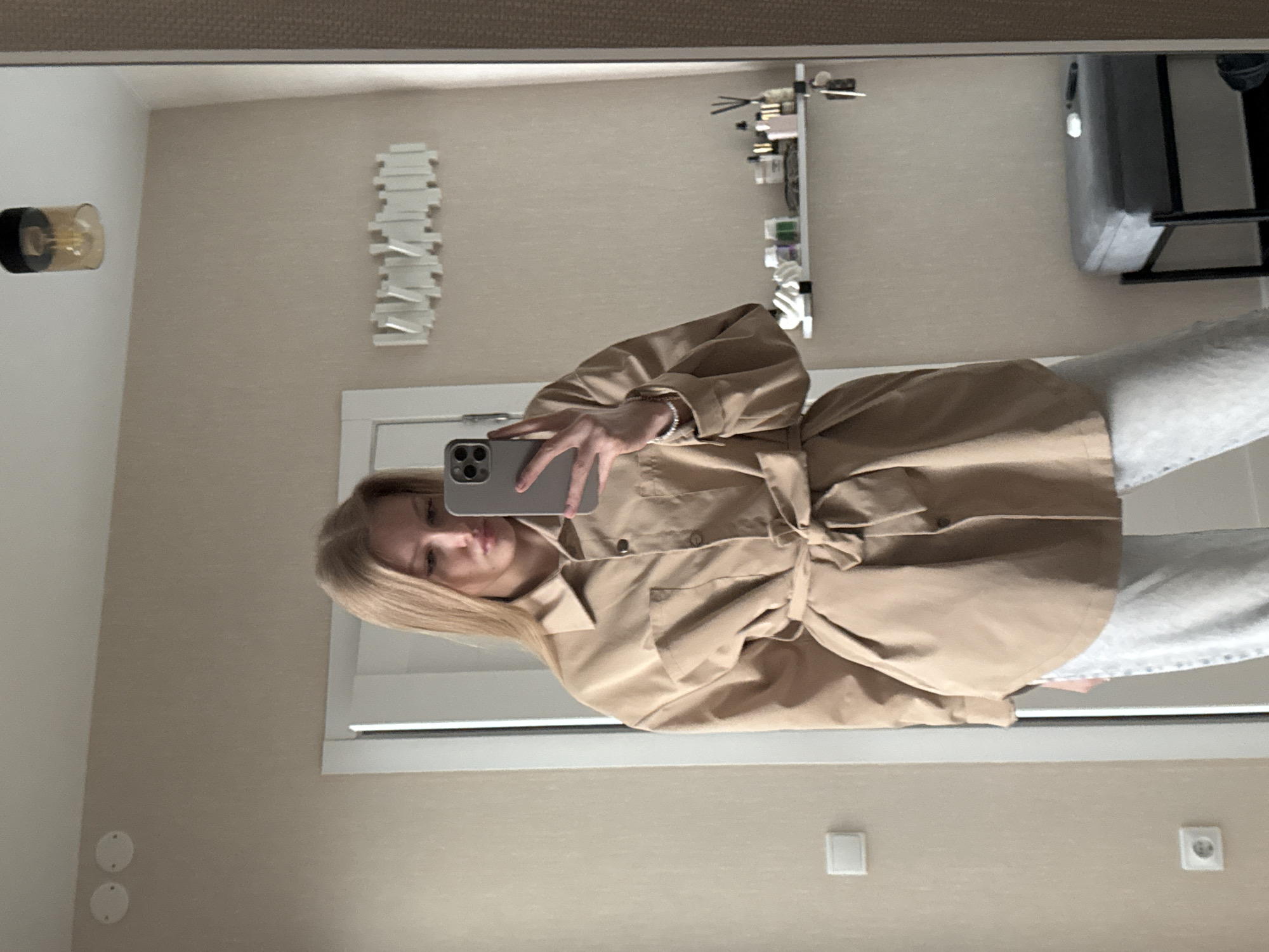 A young woman is taking a selfie in a mirror, likely in a room. She is wearing a light brown belted shirt jacket and grey sweatpants. A light fixture is visible on the wall to her left, with a decorative art piece above the door frame in the background. A small shelf with items on it and a glimpse of furniture can be seen reflected in the mirror. The environment is neutral in color. Given the location is Perm, Russia, as requested, no contextual information can be inferred from the image alone.