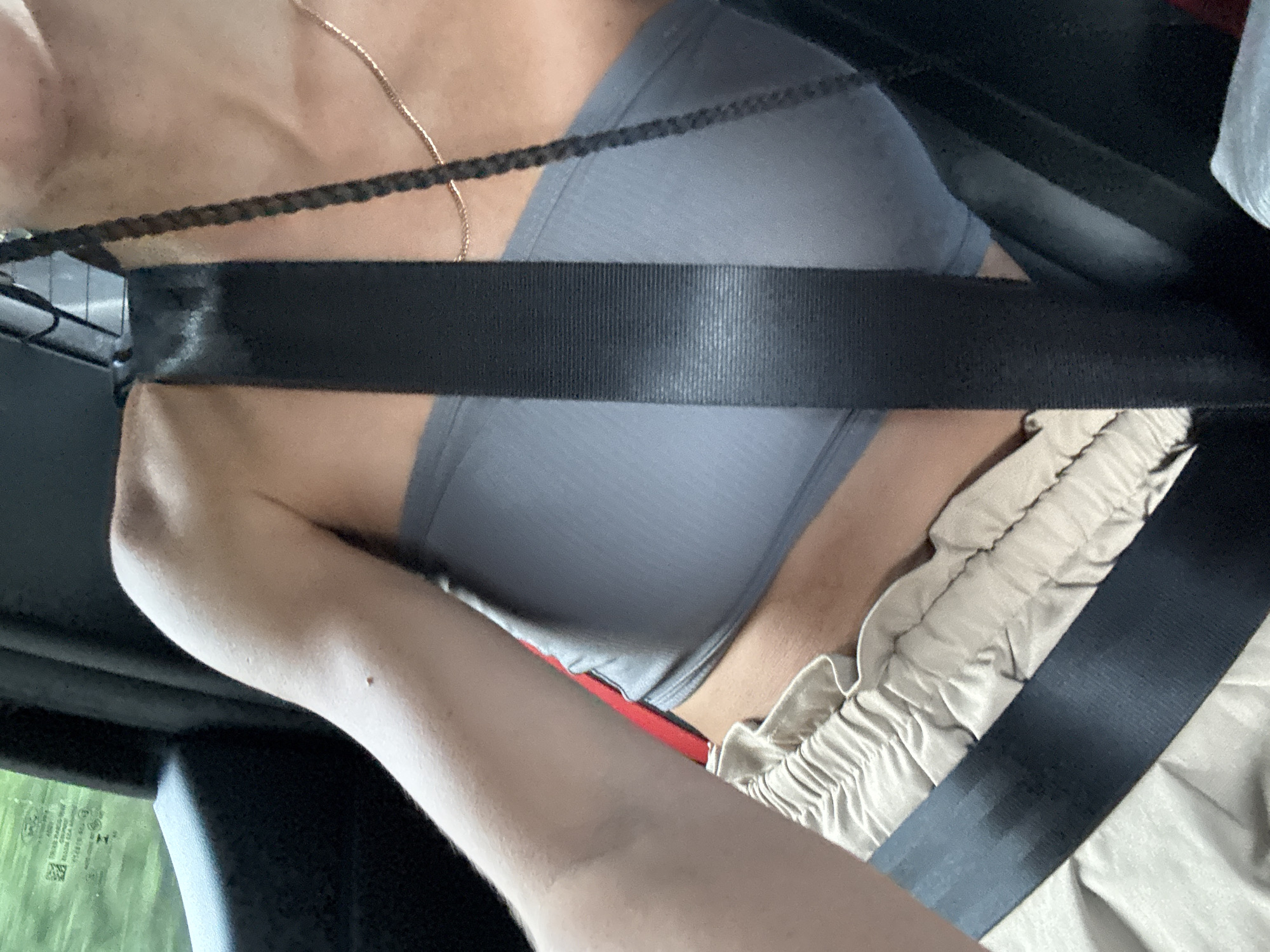 The image presents a close-up, angled view of a person seated inside a vehicle, likely a car, in Vị Xuyên Commune, Vietnam. The main subject is partially visible, showing their upper torso and right arm. A prominent black seatbelt stretches diagonally across the person's chest and horizontally across their lap, securing them.
The individual is wearing a grey top, which appears to be a form-fitting tank top or sports bra, and light-colored, possibly beige or cream-colored, baggy pants with an elastic waistband. A thin gold chain necklace is visible around their neck and collarbone. A small red band or strap peeks out from under the grey top near the waistline. The person's right arm is bent, with a small mole visible on the bicep.
Through the car window on the left, green foliage is visible, indicating it is daytime. The lighting suggests bright conditions. The scene captures the person in transit, emphasizing the use of the seatbelt. No clear, legible text is visible within the frame, though some blurry reflections or markings are present on the car window.