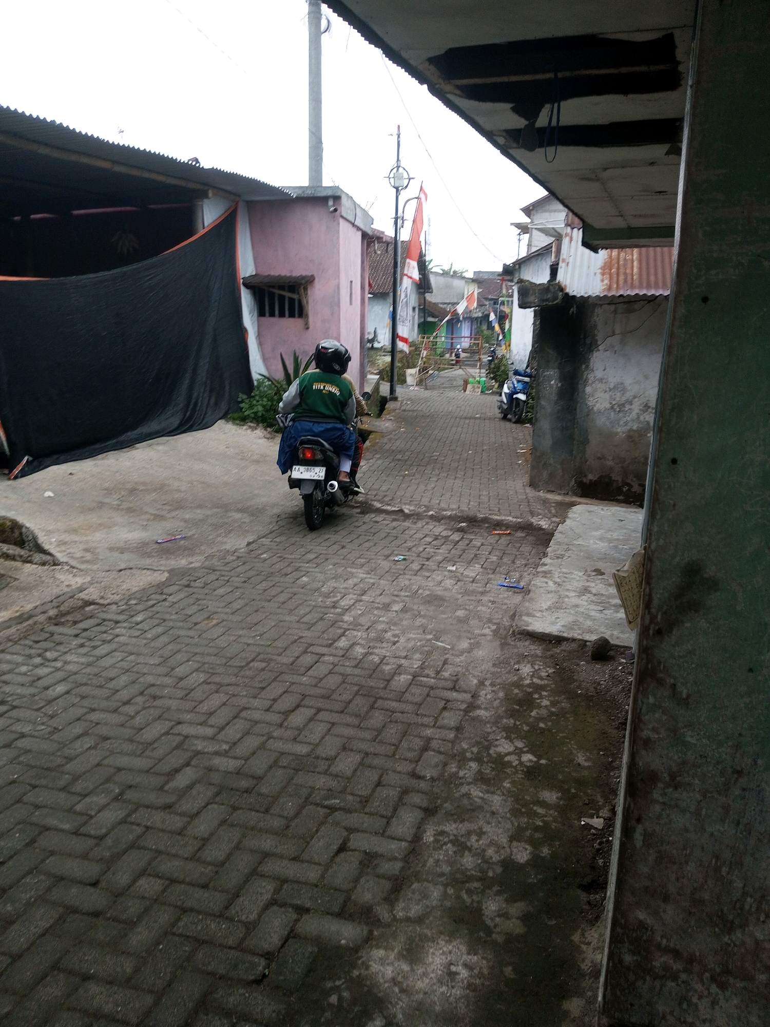 A person wearing a green jacket and a black helmet is riding a motorcycle down a narrow, paved alley in the city of Andongsili, Indonesia. The alley is lined with buildings, some with corrugated metal roofs. A black tarp hangs on the side of one building, and Indonesian flags are visible in the distance. The ground is paved with interlocking bricks. The sky is overcast.