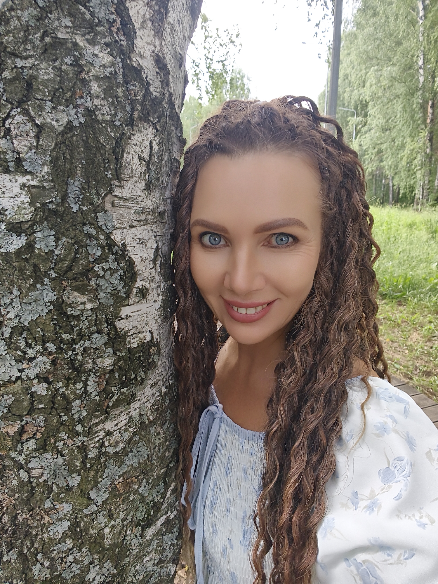 This media file is a portrait of a woman leaning against a birch tree in a park-like setting. The woman has long, curly brown hair and bright blue eyes. She is smiling and looking at the camera. She is wearing a light blue and white floral print dress. The birch tree has a rough, textured bark with patches of lichen. Behind the tree, there is a grassy area with more trees and foliage, suggesting an outdoor location during the daytime. The sunlight appears to be diffused, indicating it might be an overcast day or late afternoon. The overall atmosphere is relaxed and pleasant. The location is identified as Chaikovsky, Russia. There is no visible text in the image.