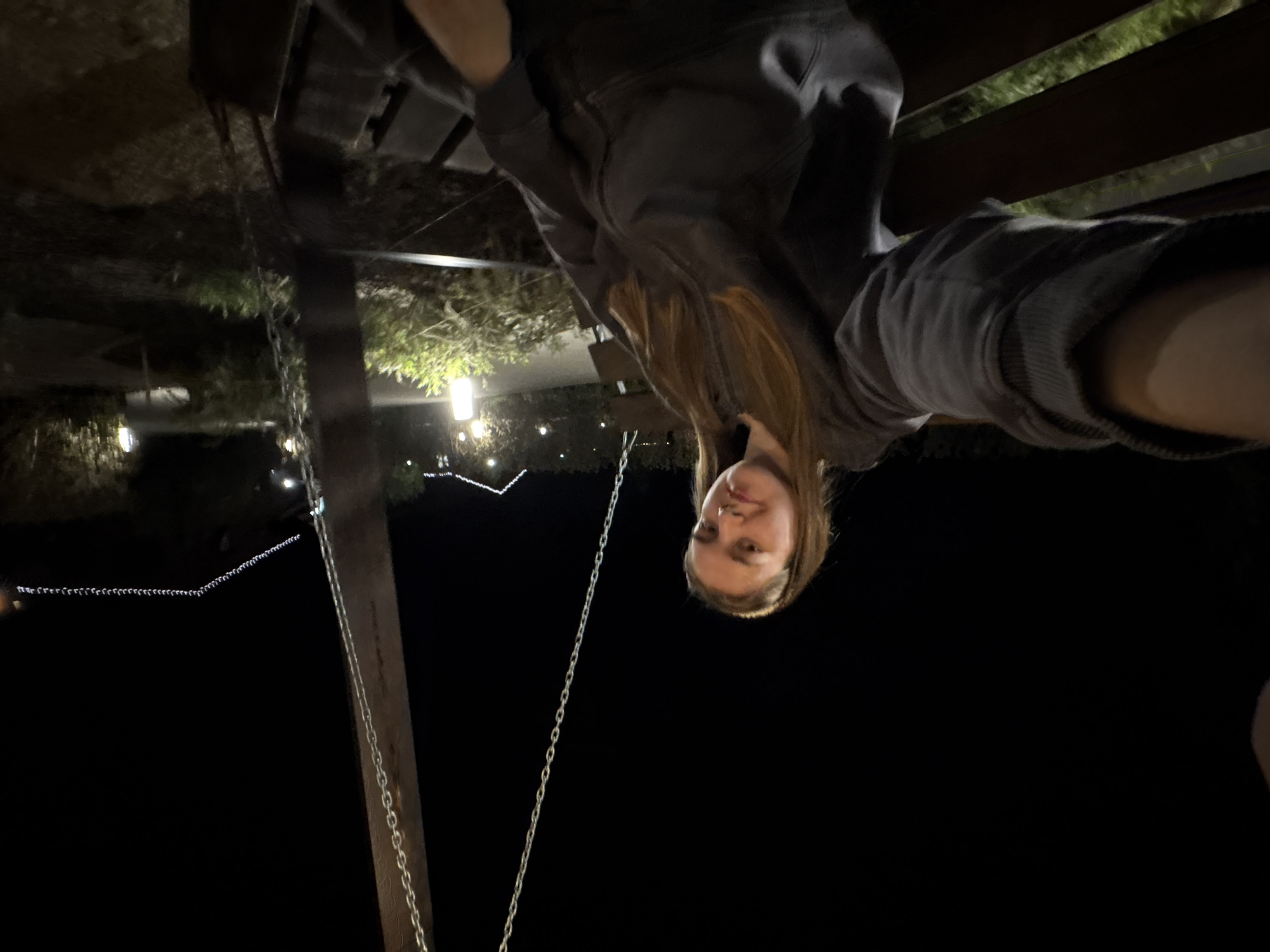 A person is sitting on a swing set at night. The background is dark with some lights visible. The person is wearing a gray hooded sweatshirt. Chains of the swing are visible. The specific location in Нижние Муллы, Russia, cannot be confirmed from the image.