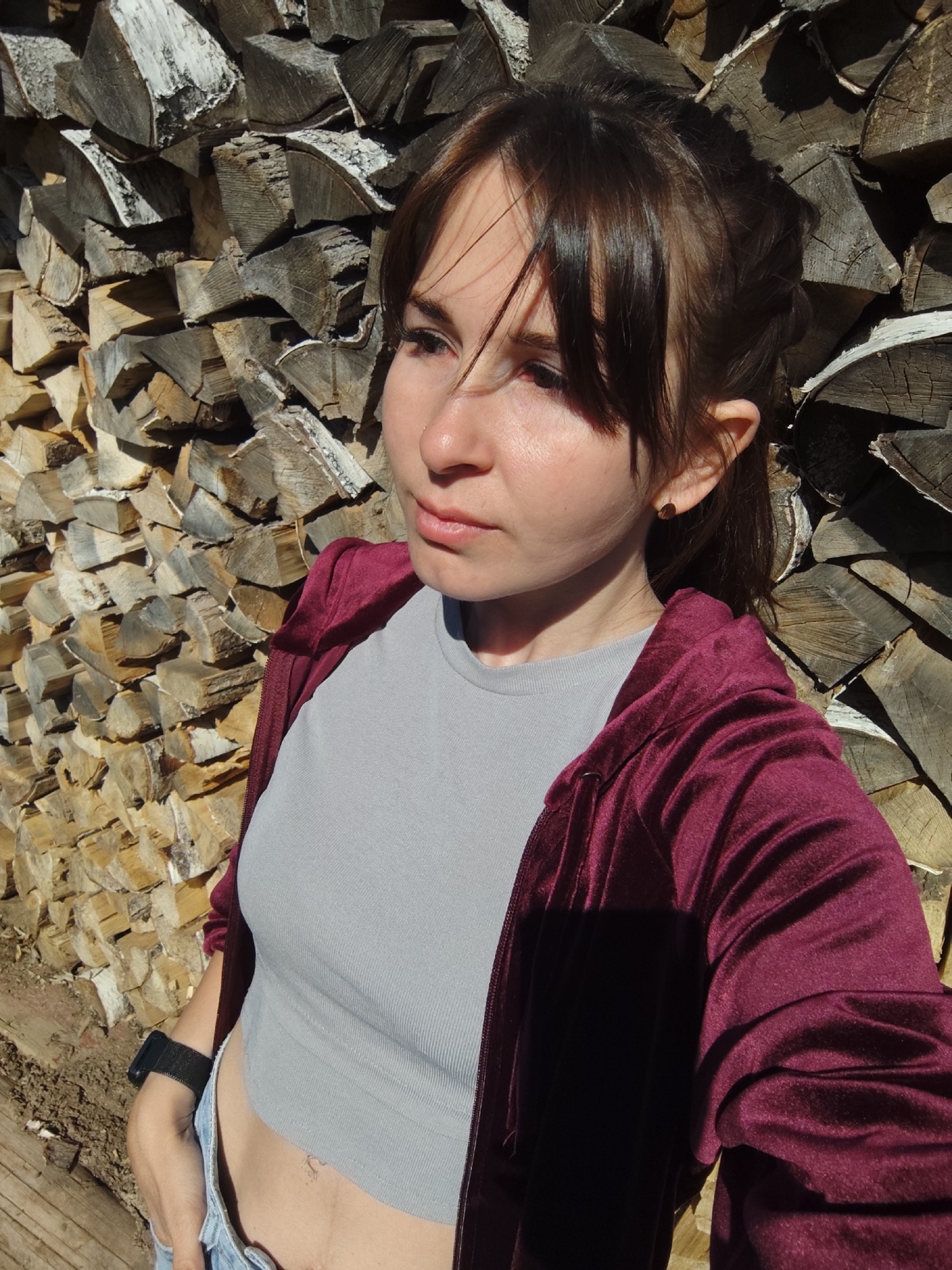 A woman stands in front of a stacked woodpile, likely in a rural or suburban area. She is fair-skinned with brown hair and bangs, styled in a casual ponytail. She wears a gray ribbed crop top, blue jeans, and a maroon velvet hoodie. Her posture suggests a relaxed pose, with one hand possibly in her pocket. A smartwatch is visible on her wrist. The woodpile is made of birch logs, with bark visible. The light suggests it is daytime, casting shadows on the wood and the woman's face. There is no evidence to support the location is in the city of Фоки, Russia.