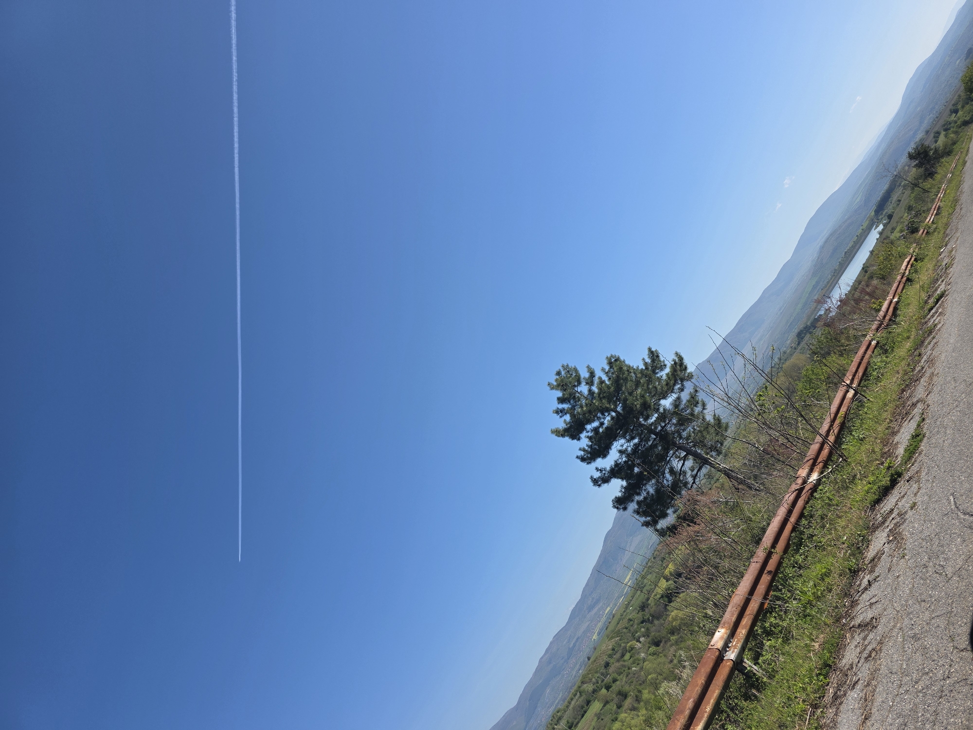 The image shows a scene in Lelintsi, Bulgaria. A blue sky with a contrail stretches above a landscape. The ground slopes downhill, with a road running along a ridge. A rusty metal guardrail separates the road from the edge. A single pine tree stands near the road, with hills and valleys visible in the distance. A body of water is also present in the distance. The photo appears to be taken during daylight hours.