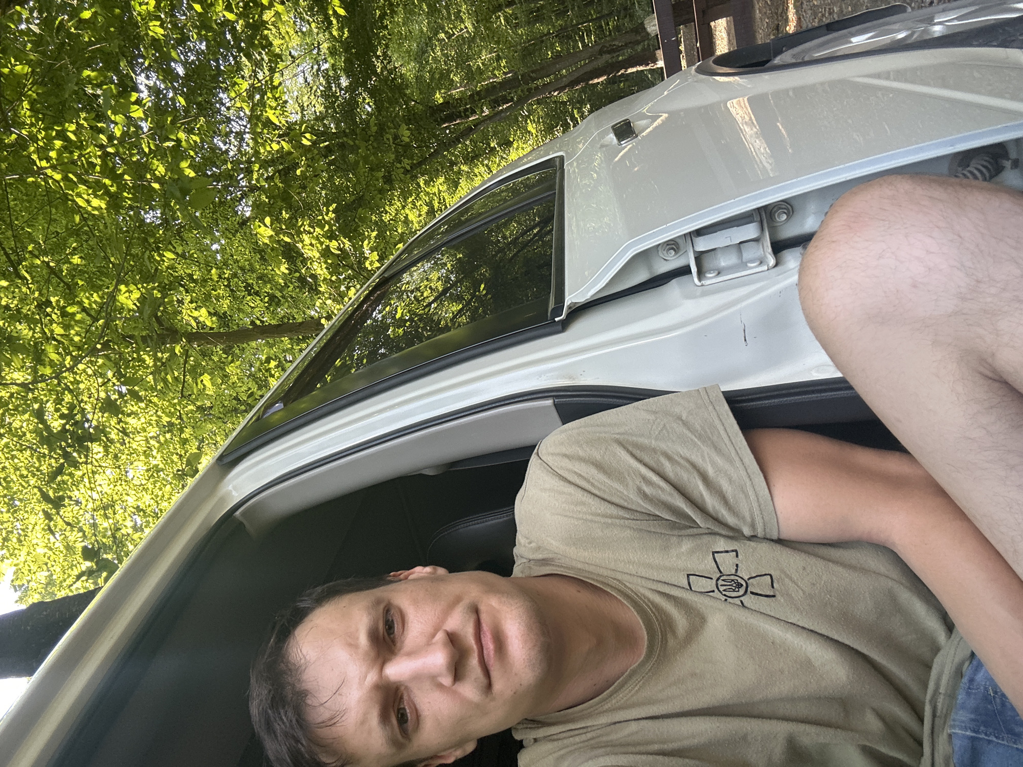 A man is pictured from the chest up, sitting in the open doorway of a white vehicle. He is looking directly at the camera with a neutral expression. He is wearing a tan t-shirt with a black emblem on the left chest. The vehicle is parked on a dirt surface, with trees and foliage forming a dense green background. The lighting suggests it is daytime. The scene appears to be in a wooded area.