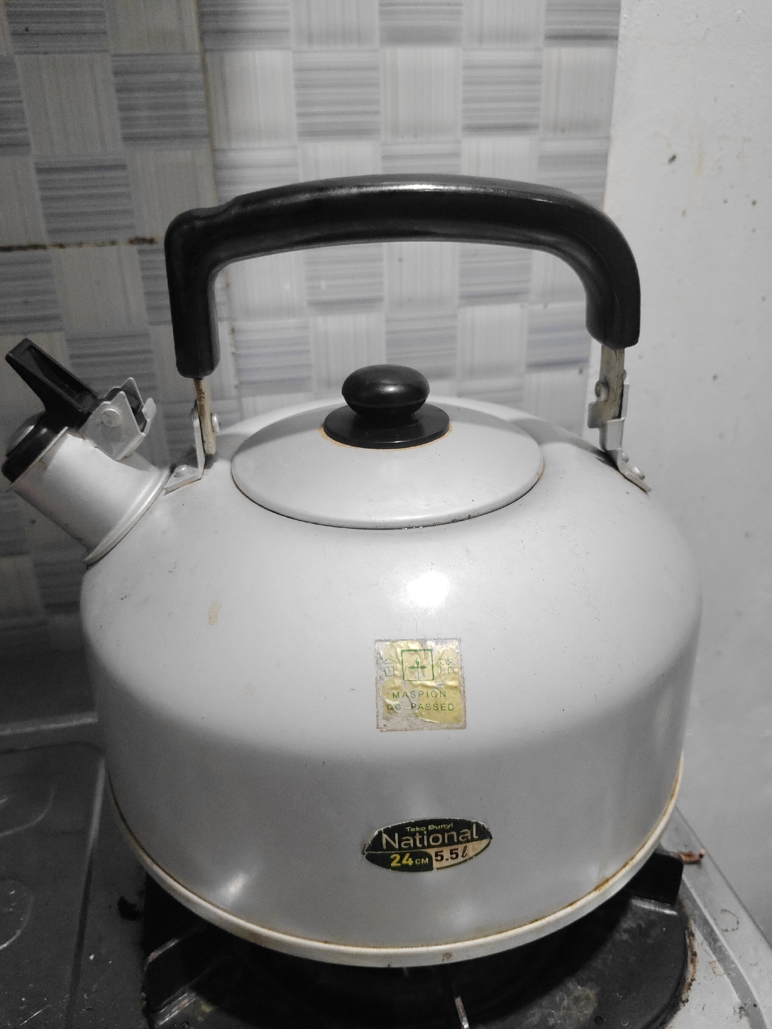 A light gray whistling kettle with a black lid and handle sits on a black gas stove burner. The kettle has a spout with a black flip-top lid. There are two labels on the side of the kettle. The upper label is rectangular and faded, with text that appears to be "MASPION QC PASSED". The lower label is oval-shaped and has "Teko Bunyi National 24 cm 5.5 L" printed on it in black and yellow text. The background consists of a tiled wall with a grid pattern of light and dark gray squares. The scene is indoors, likely in a kitchen. The city of Semambung, Indonesia is not explicitly depicted or suggested by the image itself.
