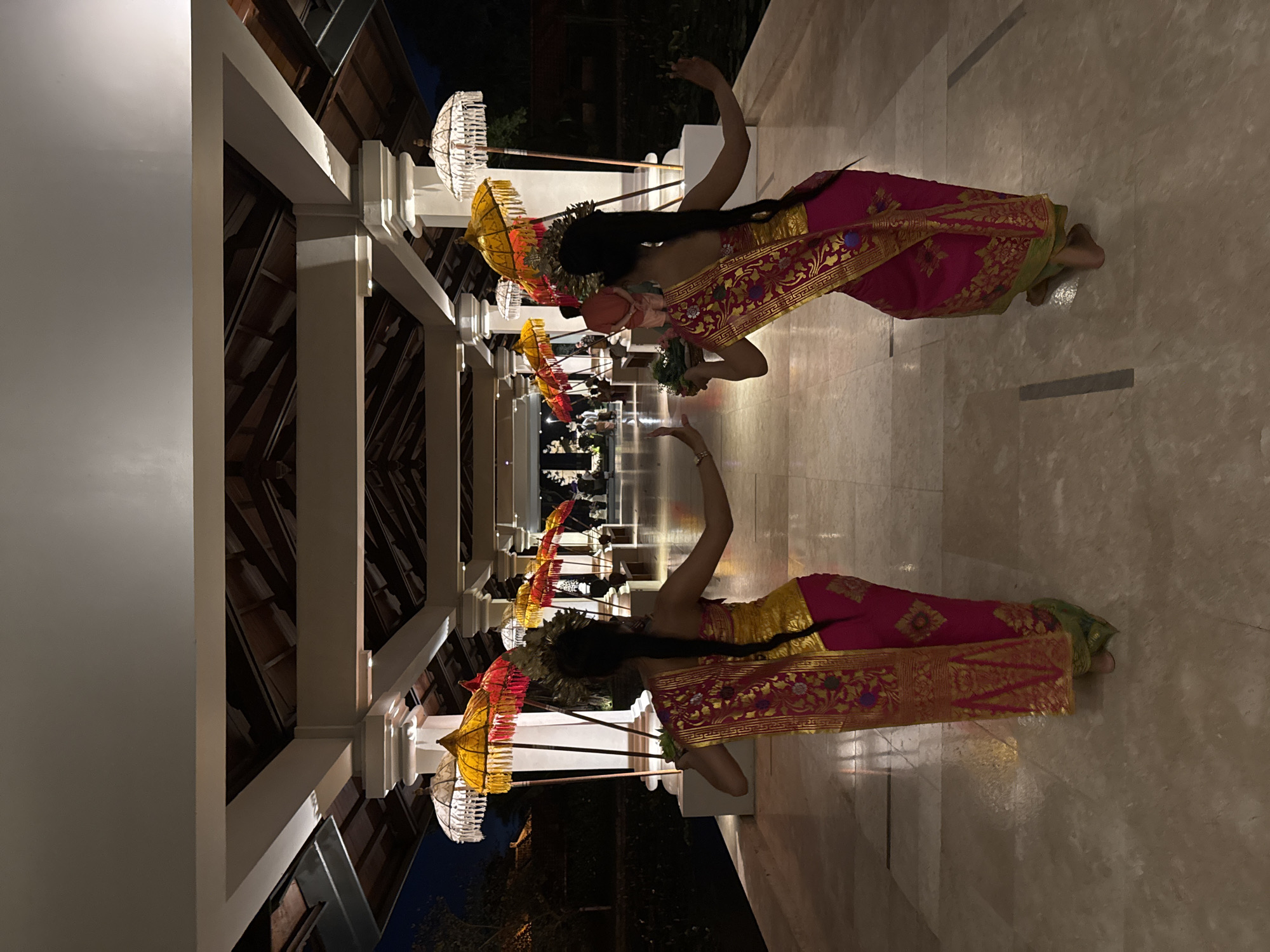 Two Balinese dancers are performing in a hallway in Kuta Selatan, Indonesia. The hallway is lined with white pillars and decorated with traditional Balinese umbrellas. The dancers are dressed in colorful costumes and headdresses, and they are holding small bowls in their hands. The floor is made of light-colored tile.