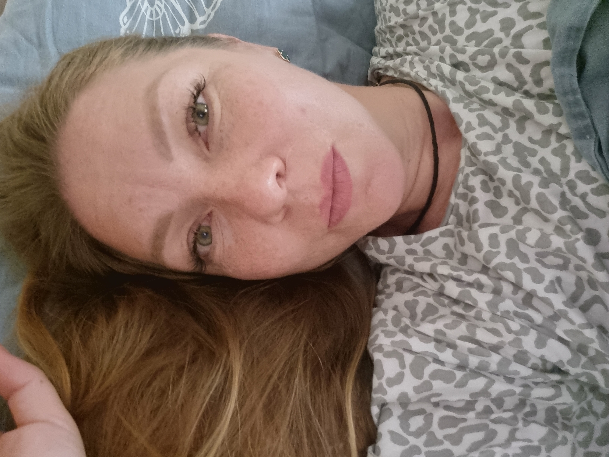 A close-up, high-angle, selfie-style shot shows a fair-skinned woman with blonde hair and green eyes lying down on a blue patterned surface. She is looking directly at the camera. Her hair is a warm blonde and falls to the left of the frame. She has fair skin with visible freckles and light pink lipstick. She is wearing a gray and white leopard-print t-shirt and a thin black choker necklace. A small, dark-colored earring is visible in her right ear. The background appears to be a bed with a blue textured pillowcase. The woman's left arm is visible in the upper left corner, raised above her head. The image has a slightly soft focus. There is no indication of the city of Chaikovsky, Russia, or any specific location.